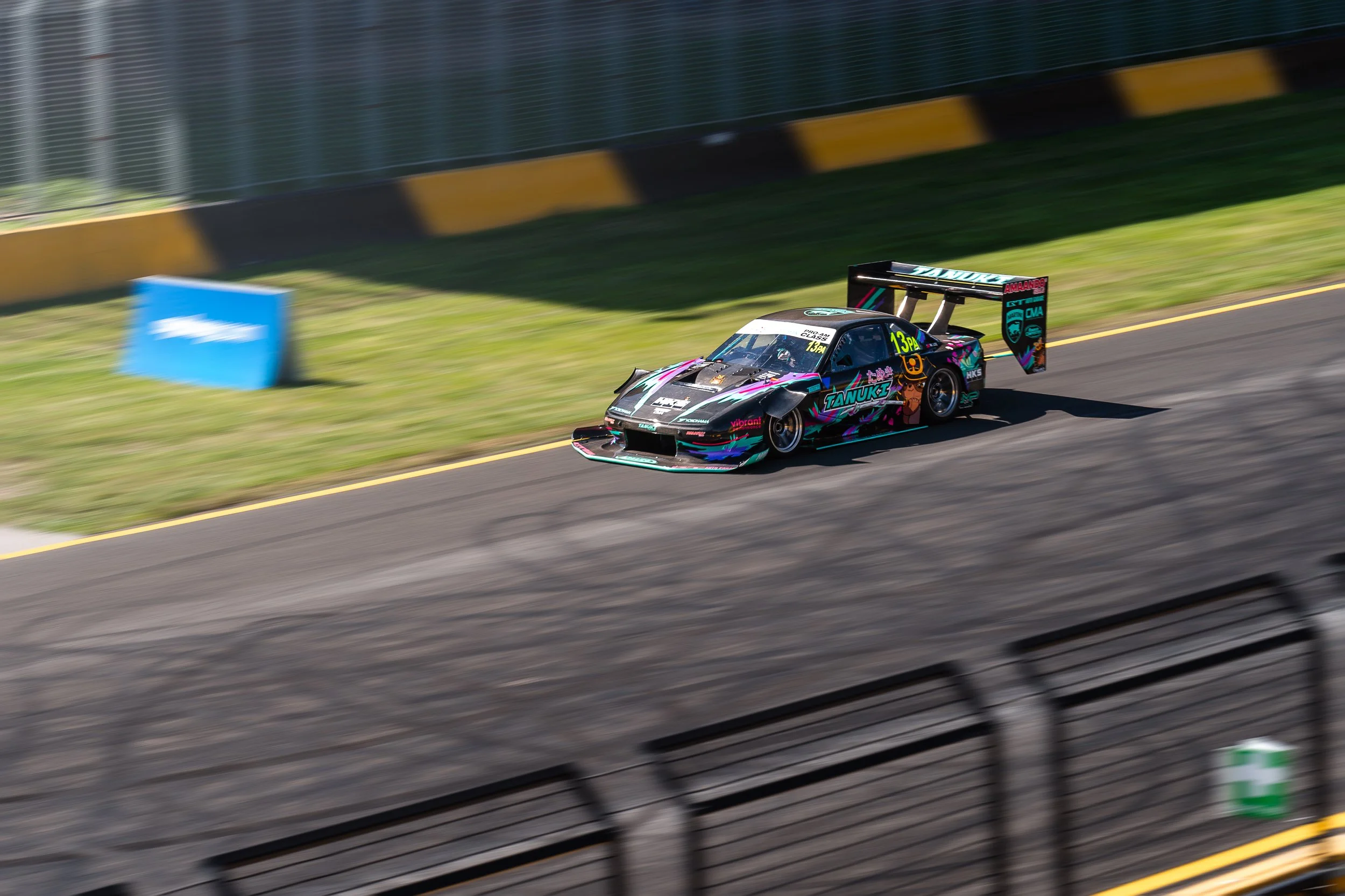 Race car speeding on track with a colorful, detailed exterior, logo, and large rear wing, blurred background and fences.