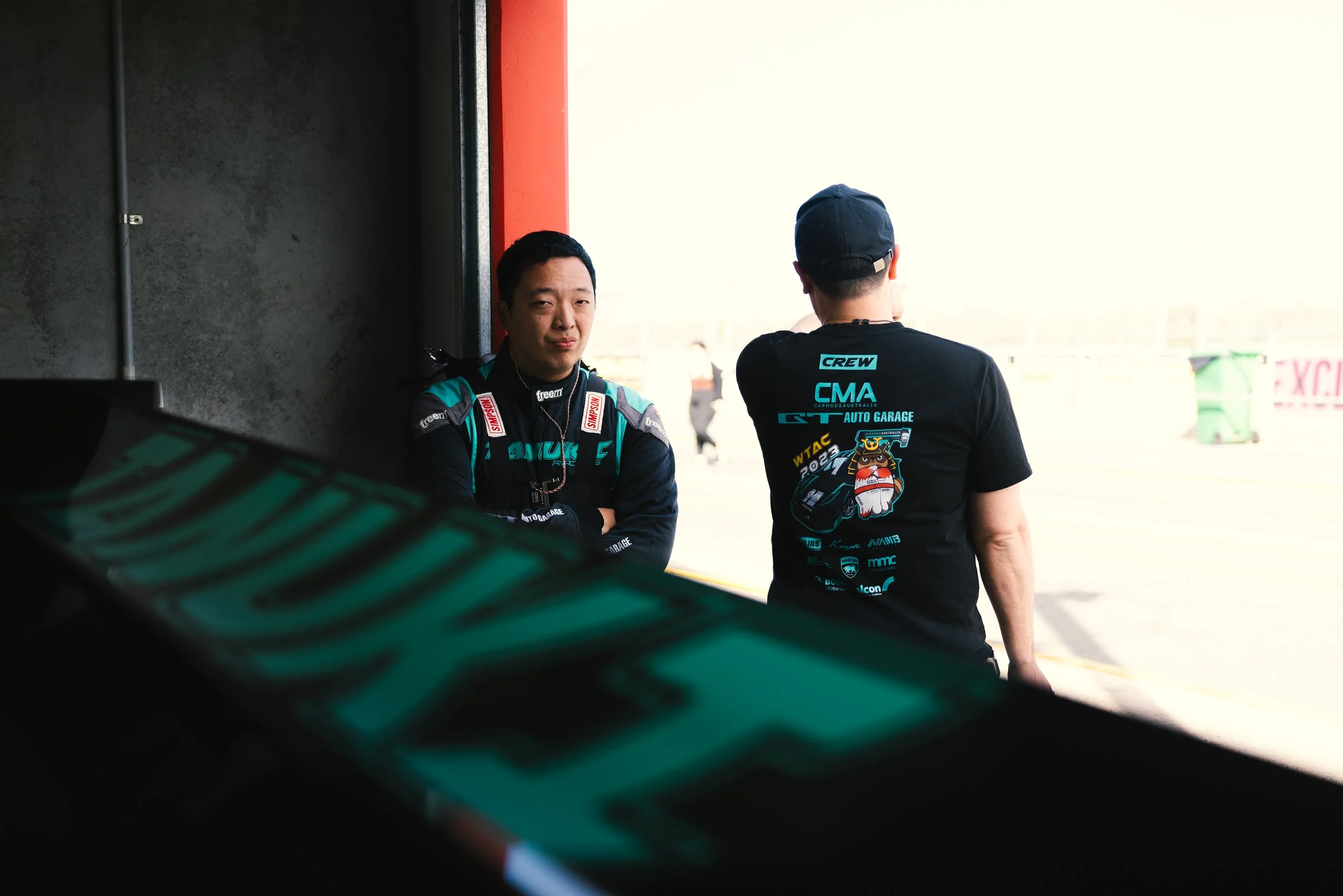Two race car drivers standing near the entrance of a garage, one facing the camera with arms crossed and the other with his back turned, wearing racing suits and casual clothes.