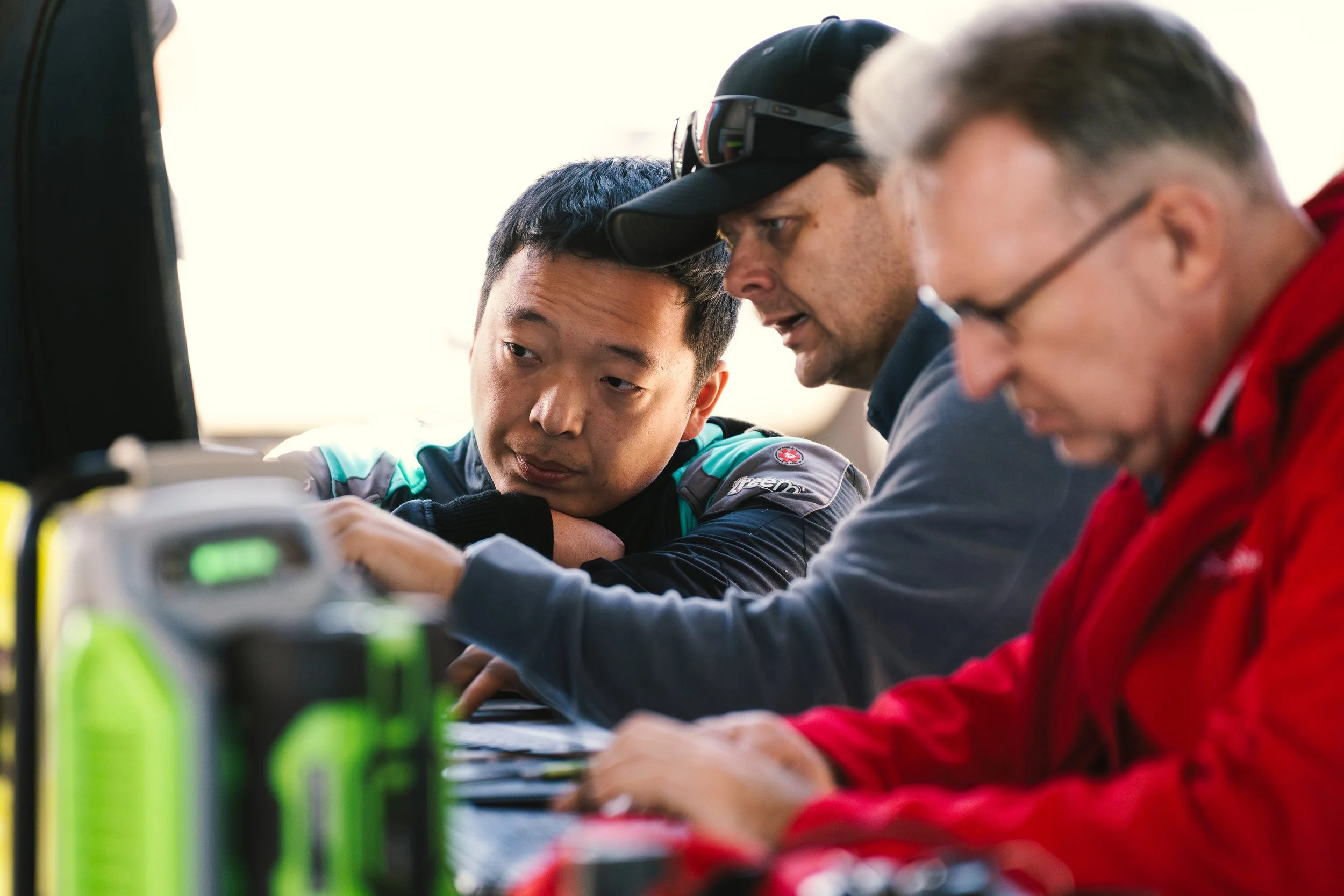 Three men are closely inspecting a computer screen at a table, appearing focused and engaged in a discussion.