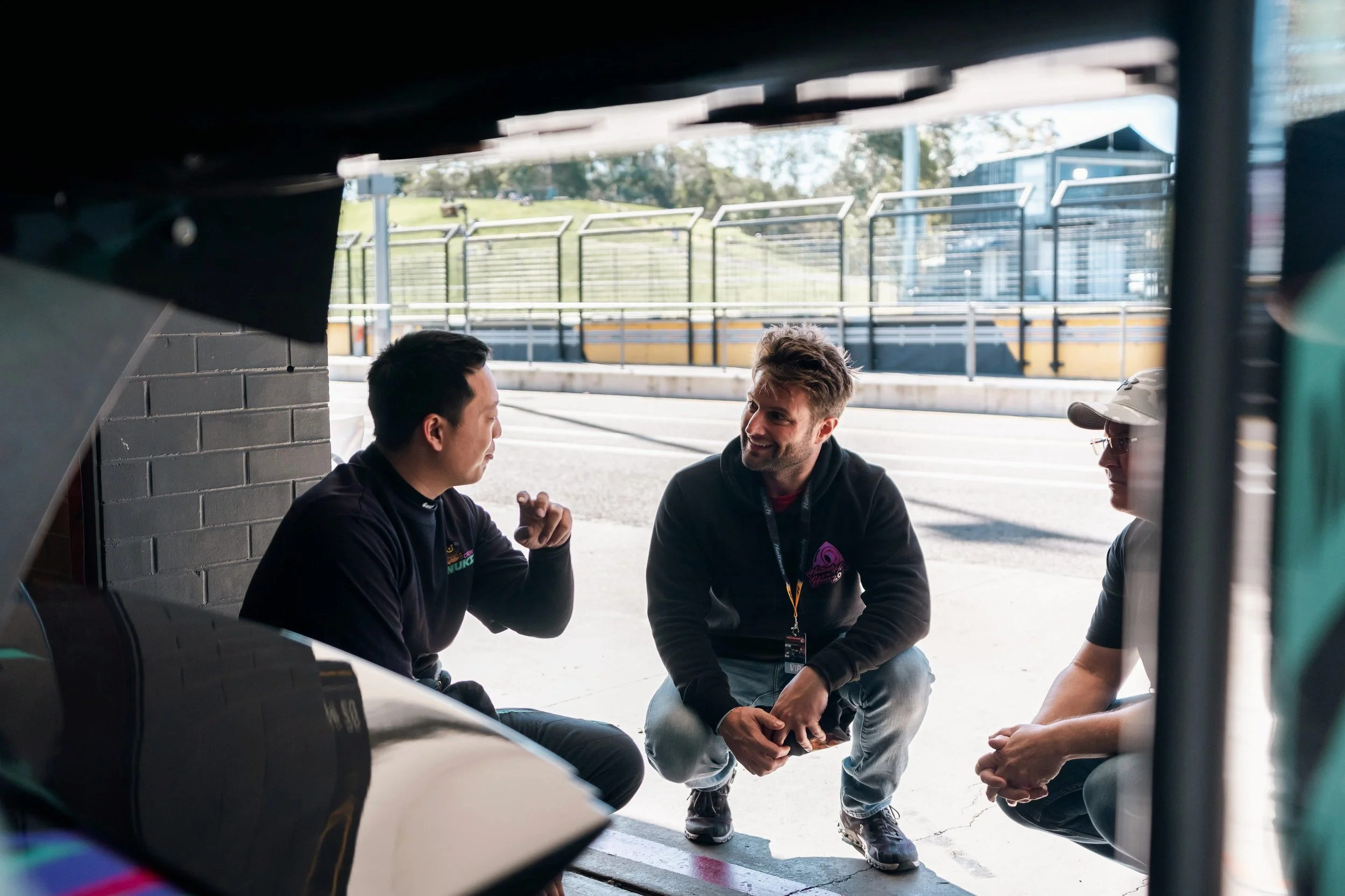 Three men having a conversation under a race car, with a garage and racetrack in the background.