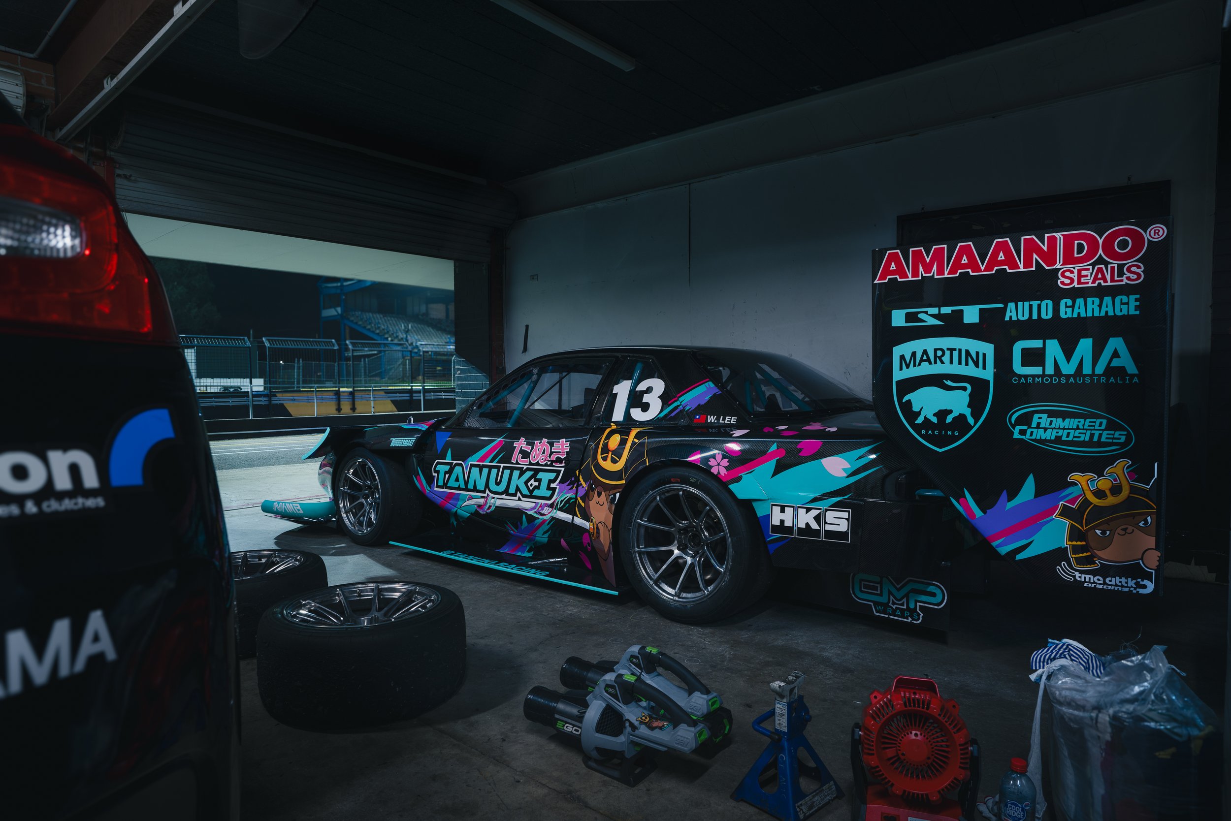 A race car with vibrant graphics and sponsorship decals parked in a garage with tools and spare tires nearby.