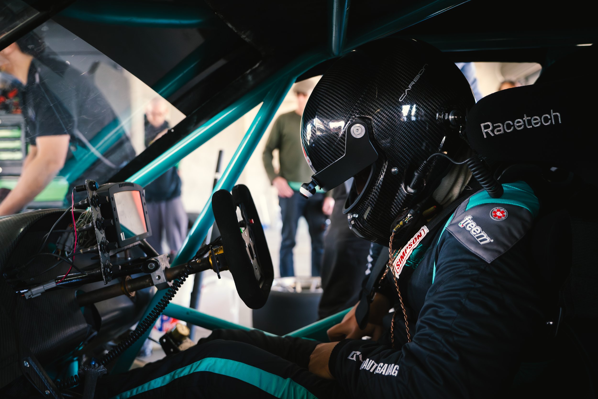 A race car driver wearing a black carbon fiber helmet and racing suit sitting inside a race car, with a digital display and racing equipment visible inside the cockpit, and several people standing outside the vehicle in the background.