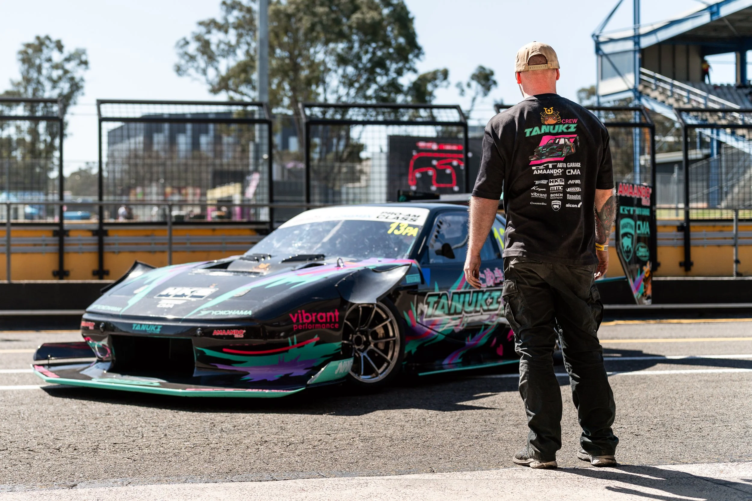 A person standing near a race car on a racetrack, with trees and fenced spectator area in the background.