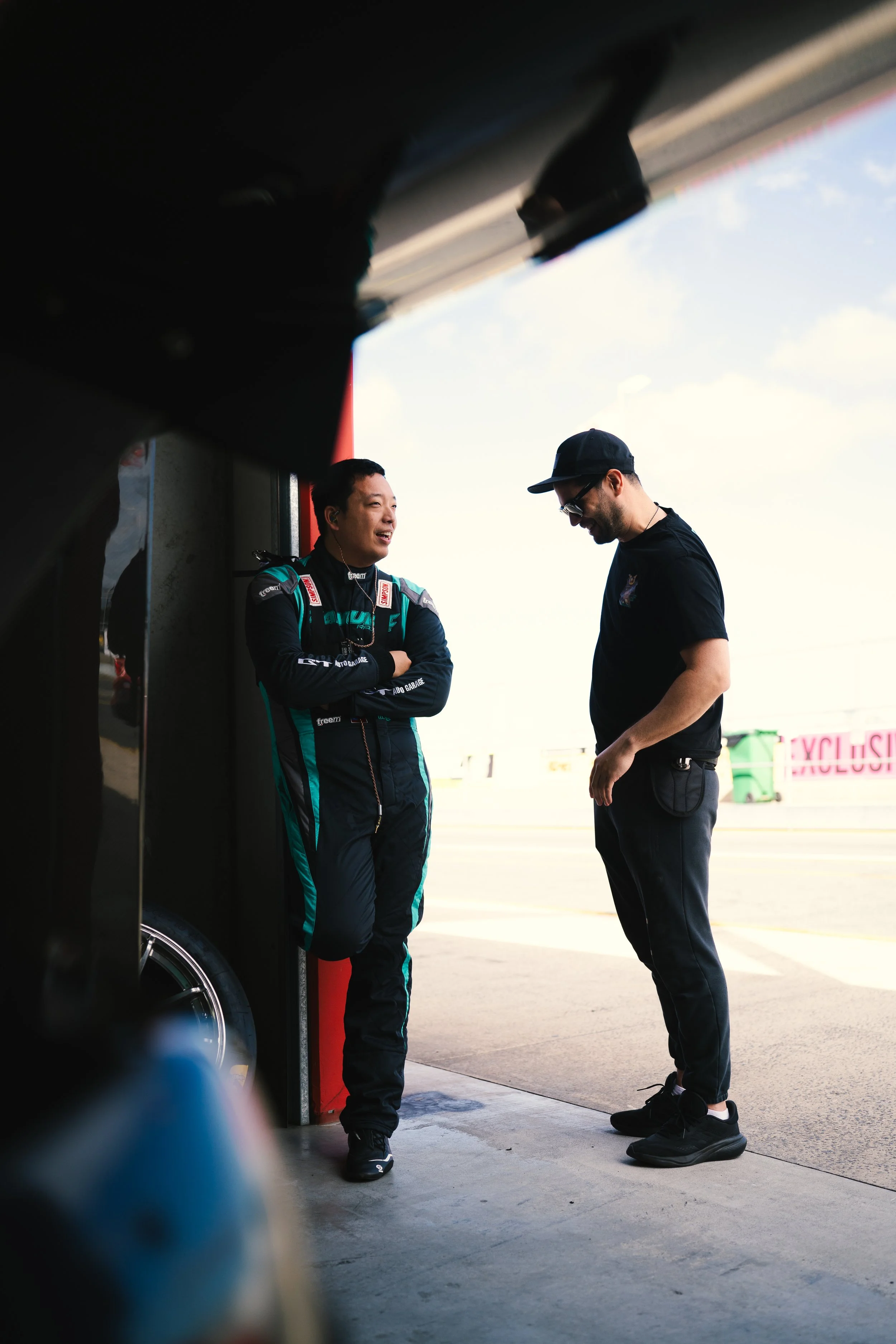 A race car driver and an official standing and talking in a garage area at a race track, with a bright sky in the background.