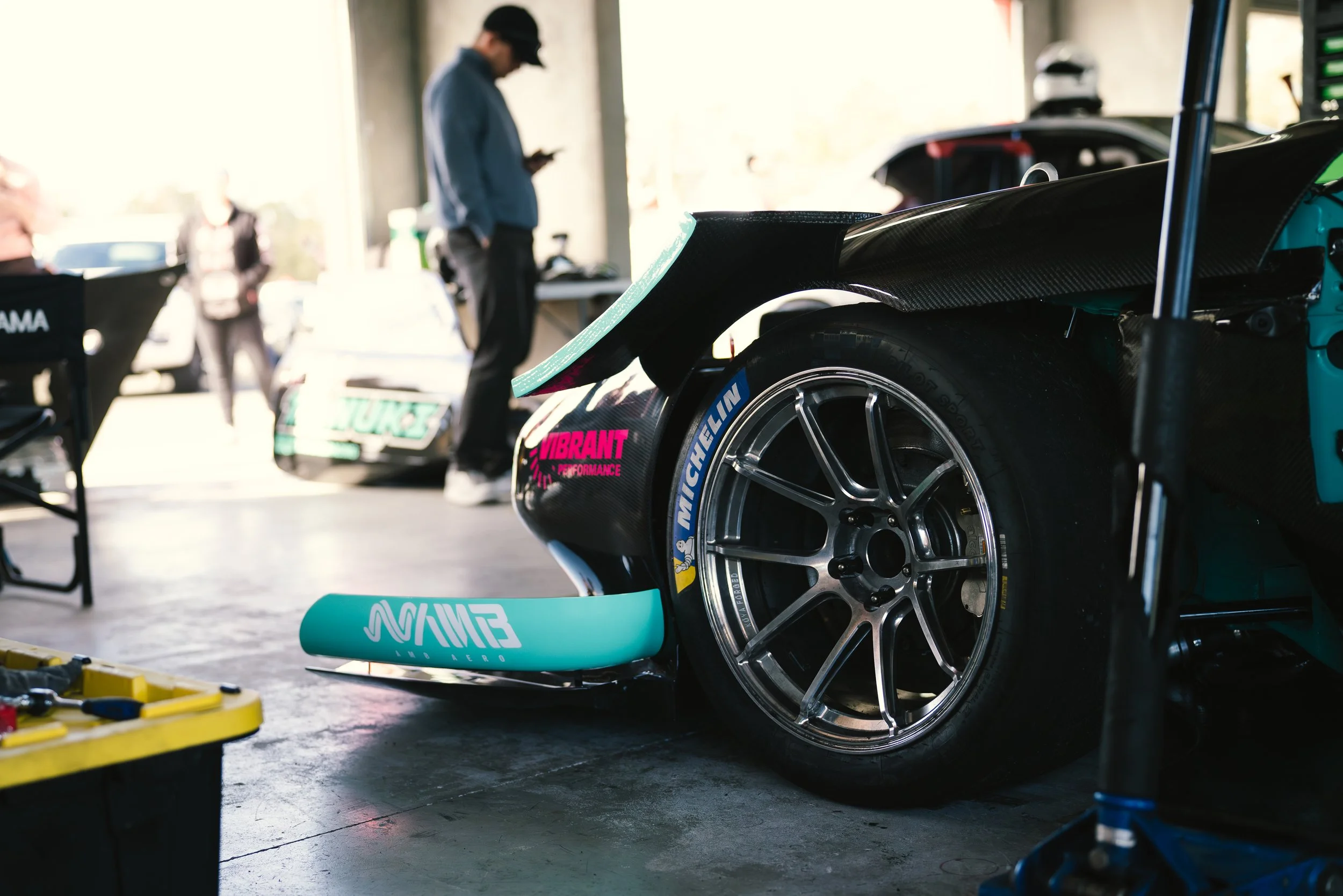 Close-up of a race car wheel and rear wing in a garage, with mechanics and equipment in the background.