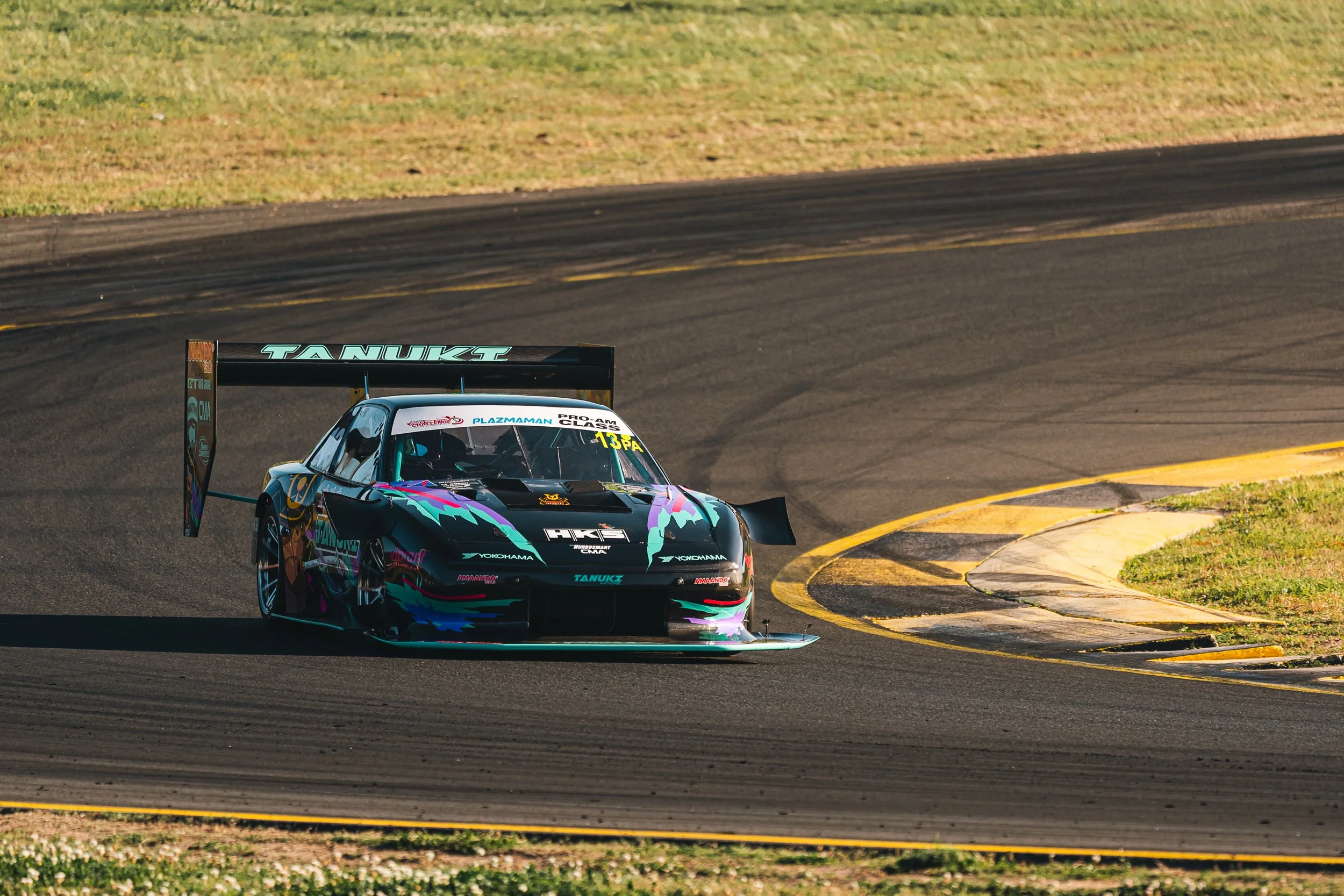 A race car with a colorful, abstract paint job navigating a curve on a racetrack, with grass and tire barriers in the background.