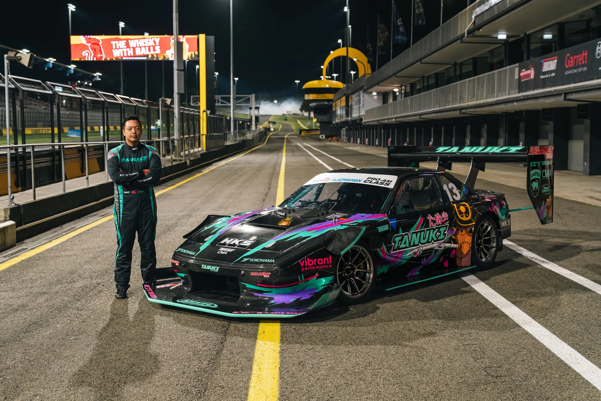 A racing driver standing confidently beside a custom race car in a nighttime pit lane with empty grandstand and enclosed garages. The car is decorated with vibrant colorful graphics, sponsor logos, and a large rear wing.