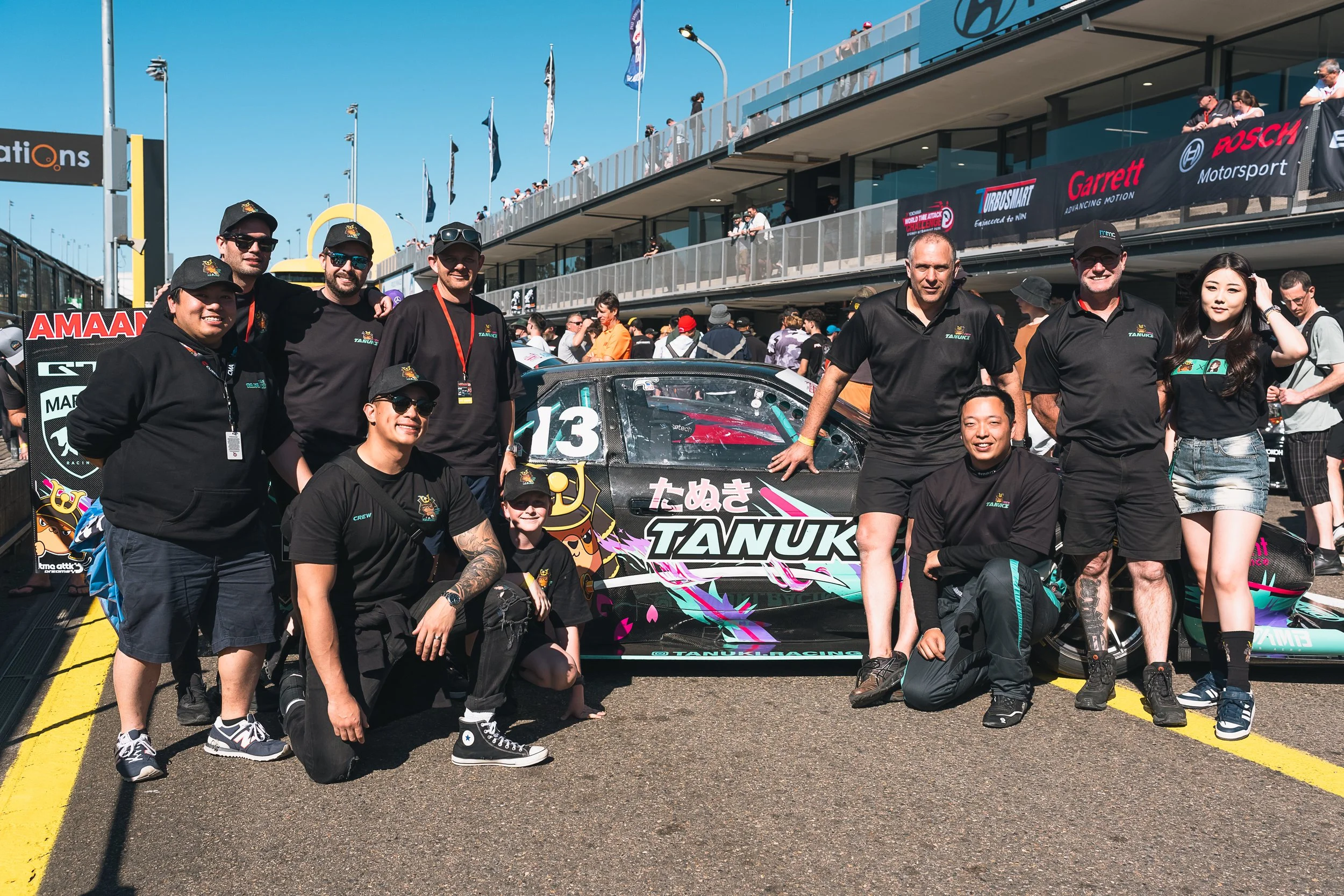 Group of people smiling and posing in front of a decorated race car with the number 13, at a motorsport event, with spectators and a pit building in the background.