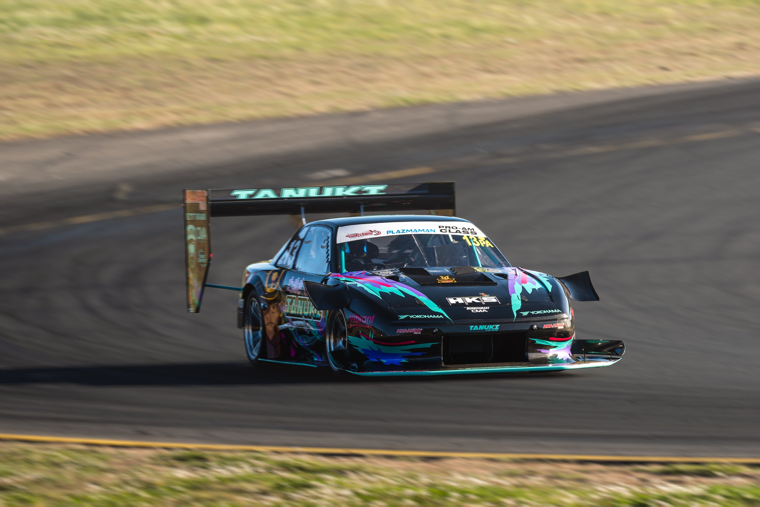 A race car moving on a racetrack during daytime, featuring a black, teal, purple, and pink color scheme, with a large rear wing and various sponsor logos.
