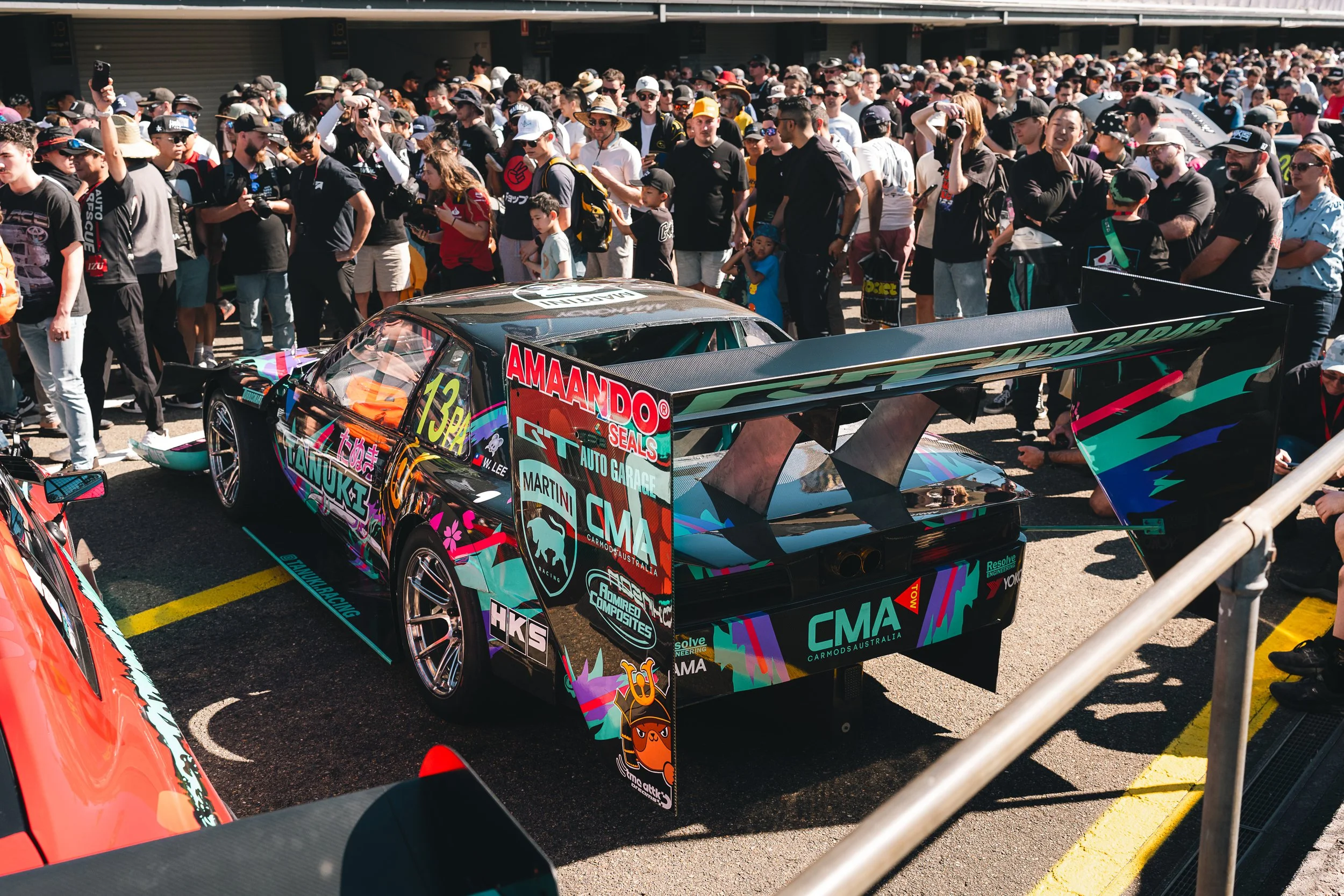 A race car with colorful artwork and sponsorship labels on it parked in a crowded paddock area during a racing event.