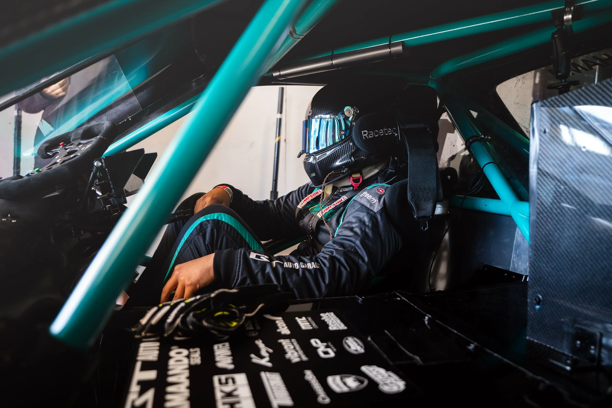 Race car driver in racing suit and helmet sitting inside a race car with safety harnesses.