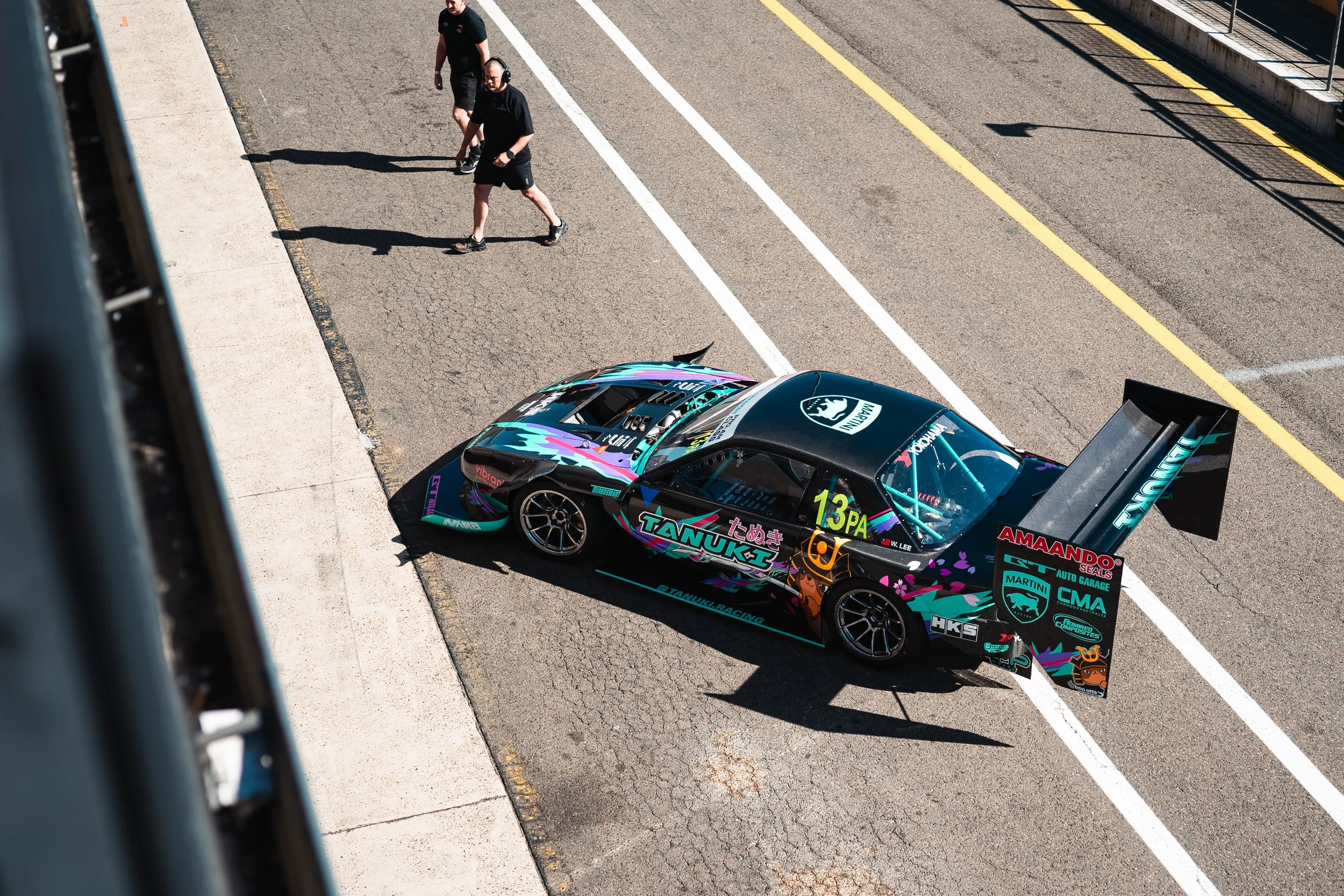 High-angle view of a colorful racing car with a large rear wing in the pit lane, with two crew members walking nearby on the asphalt surface with white and yellow lines.
