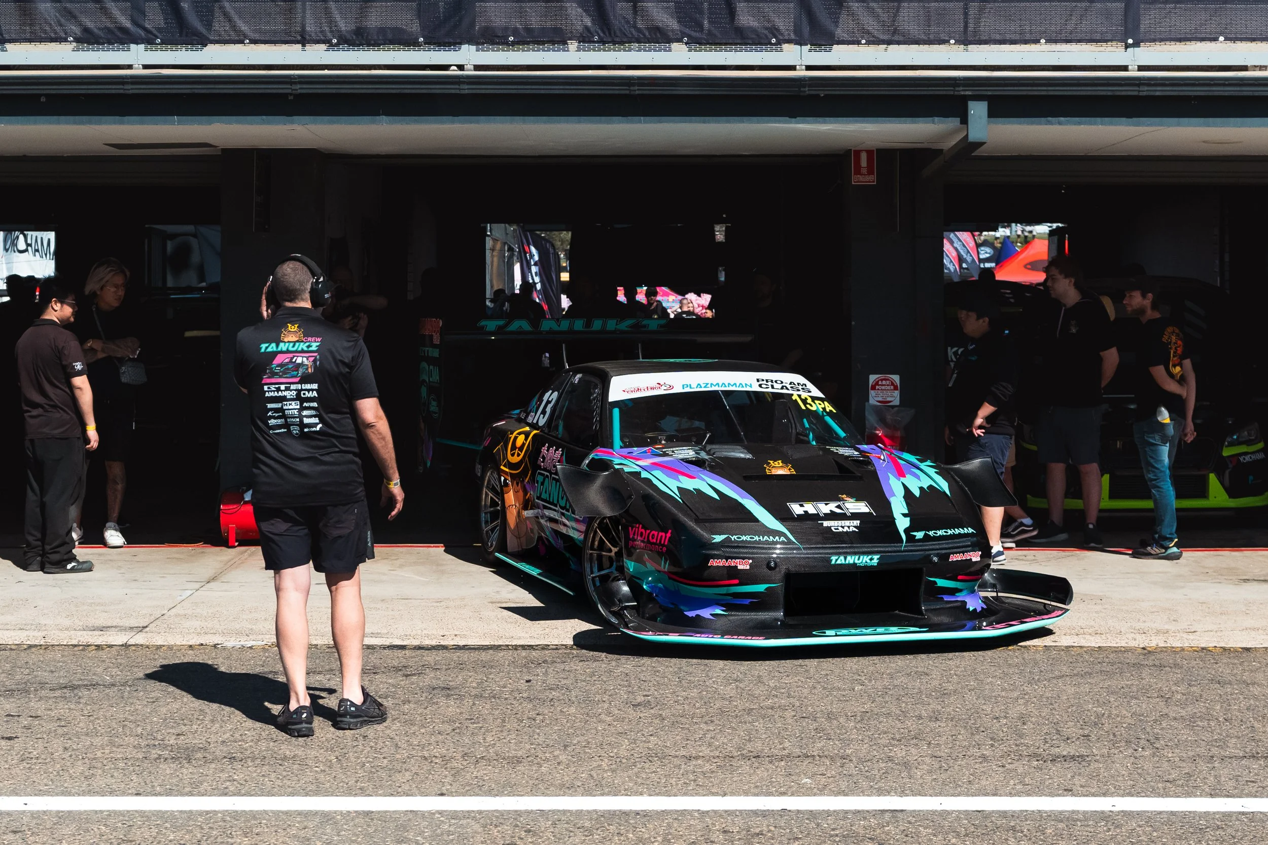 Race car in pit garage with team members around, colorful livery with branding, and a person wearing a headset observing the scene.