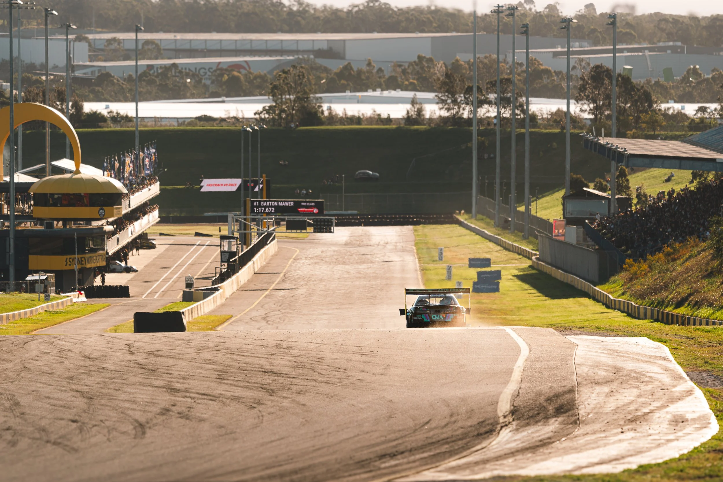 A race car on a racing track during a motorsport event, with grandstands and digital boards in the background.