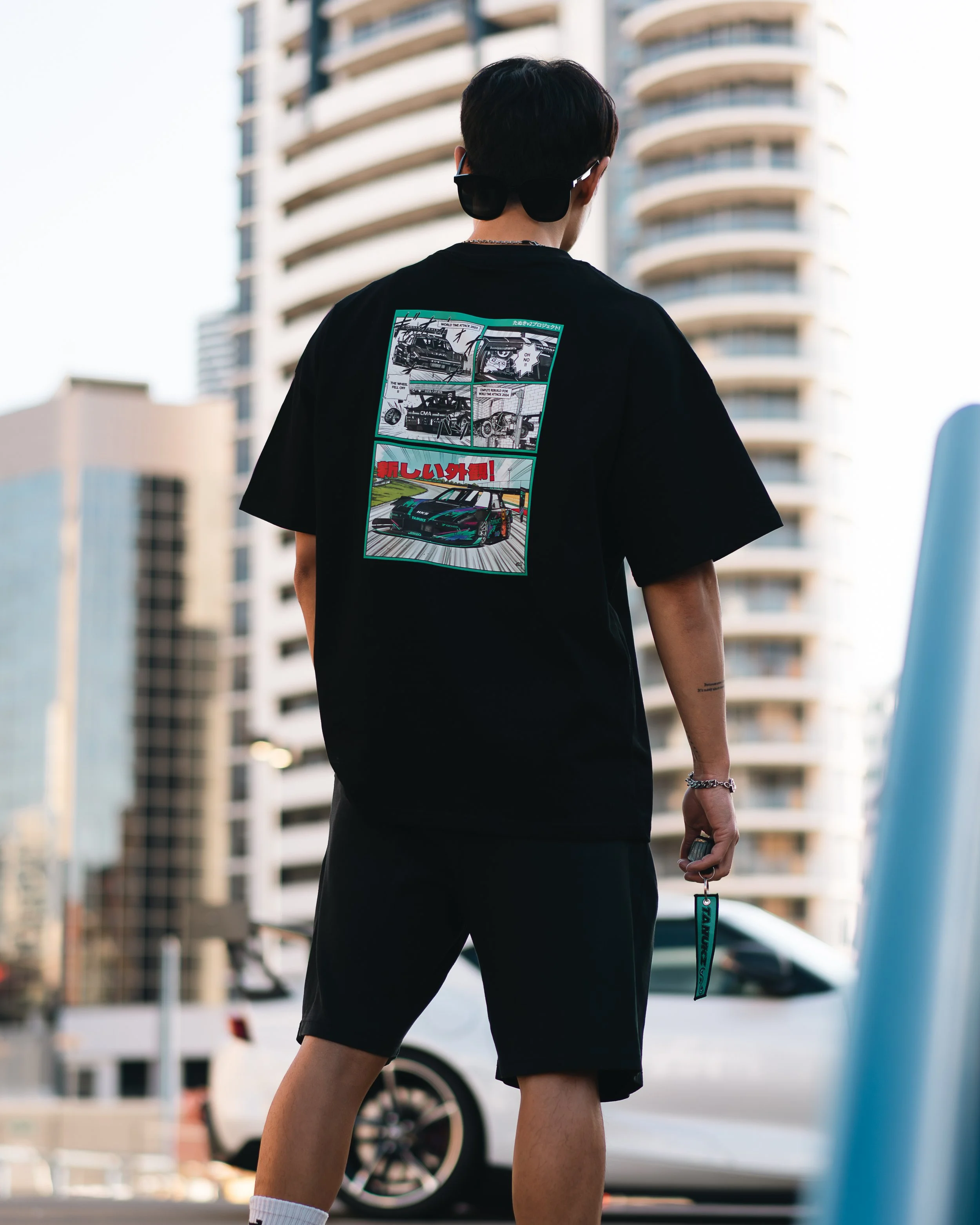 Person wearing black t-shirt with graphic print of cars, standing outdoors in an urban setting with tall buildings.