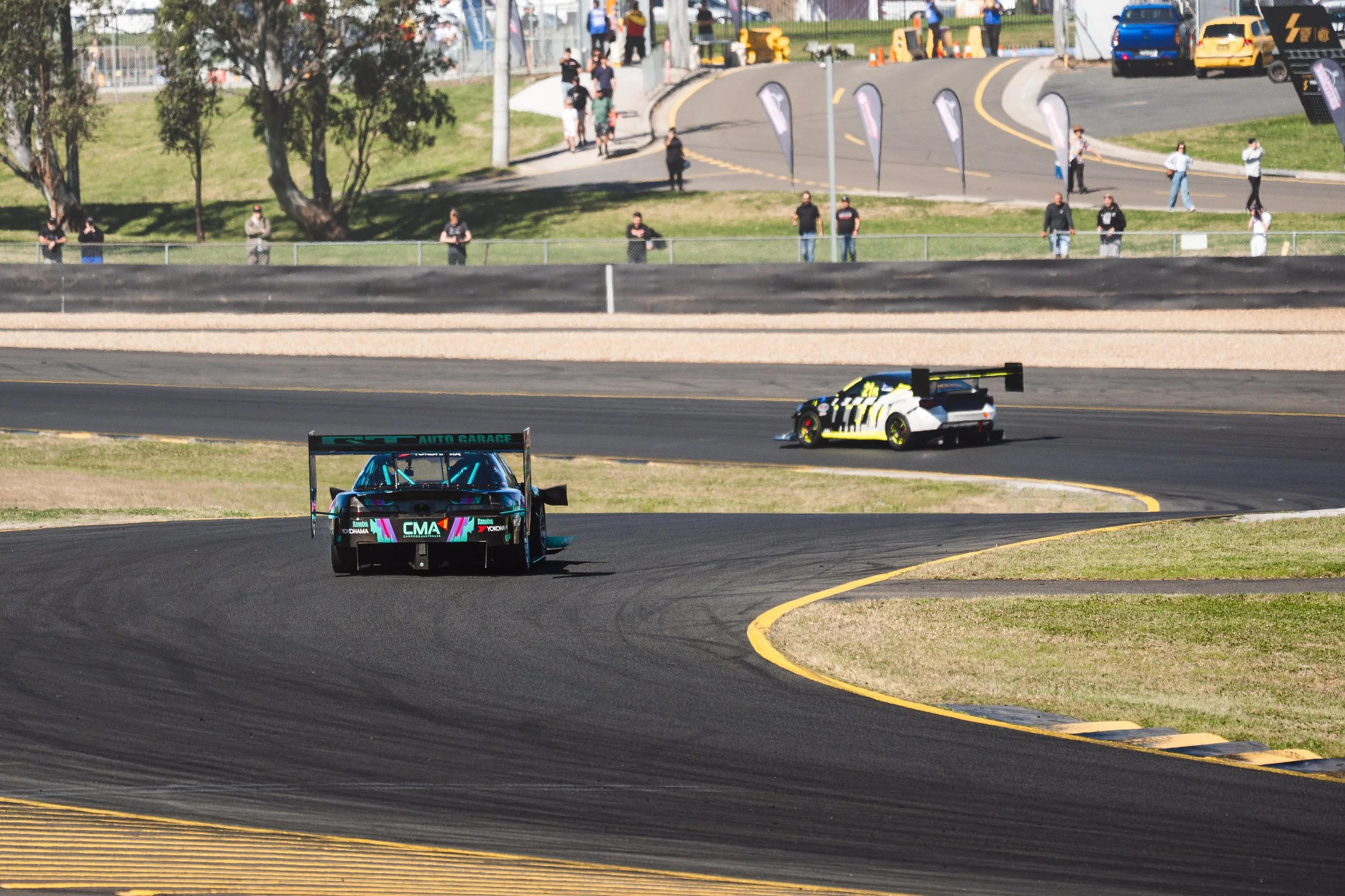 Two race cars navigate a curve on a race track, with spectators watching from the grassy area behind barriers in the background.