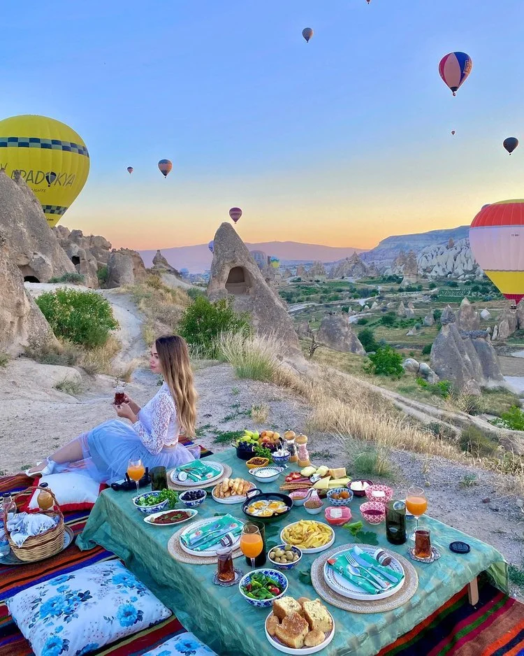 A beautifully arranged breakfast table set against the backdrop of Cappadocia's stunning landscape at sunrise, featuring a variety of traditional Turkish dishes such as olives, cheeses, fresh bread, and honey. The warm golden light of the rising sun