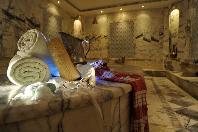 A tranquil scene inside a traditional Cappadocia Turkish Bath (Hamam), with soft lighting reflecting off the marble floors and walls. Steam fills the air as a person relaxes on a warm stone slab, preparing for a cleansing ritual. The ornate details a