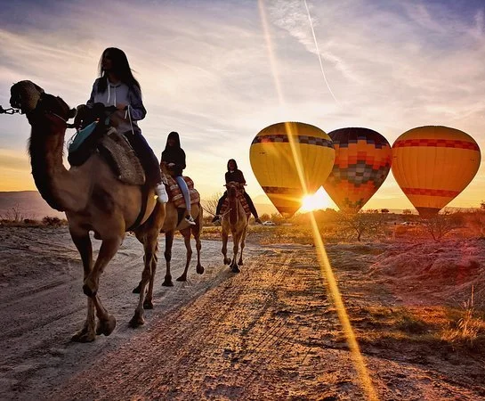 A group of tourists on a camel safari in Cappadocia, moving through a vast desert-like landscape with iconic fairy chimneys and rock formations in the background. The camels are adorned with traditional saddles, and the riders are enjoying a unique v