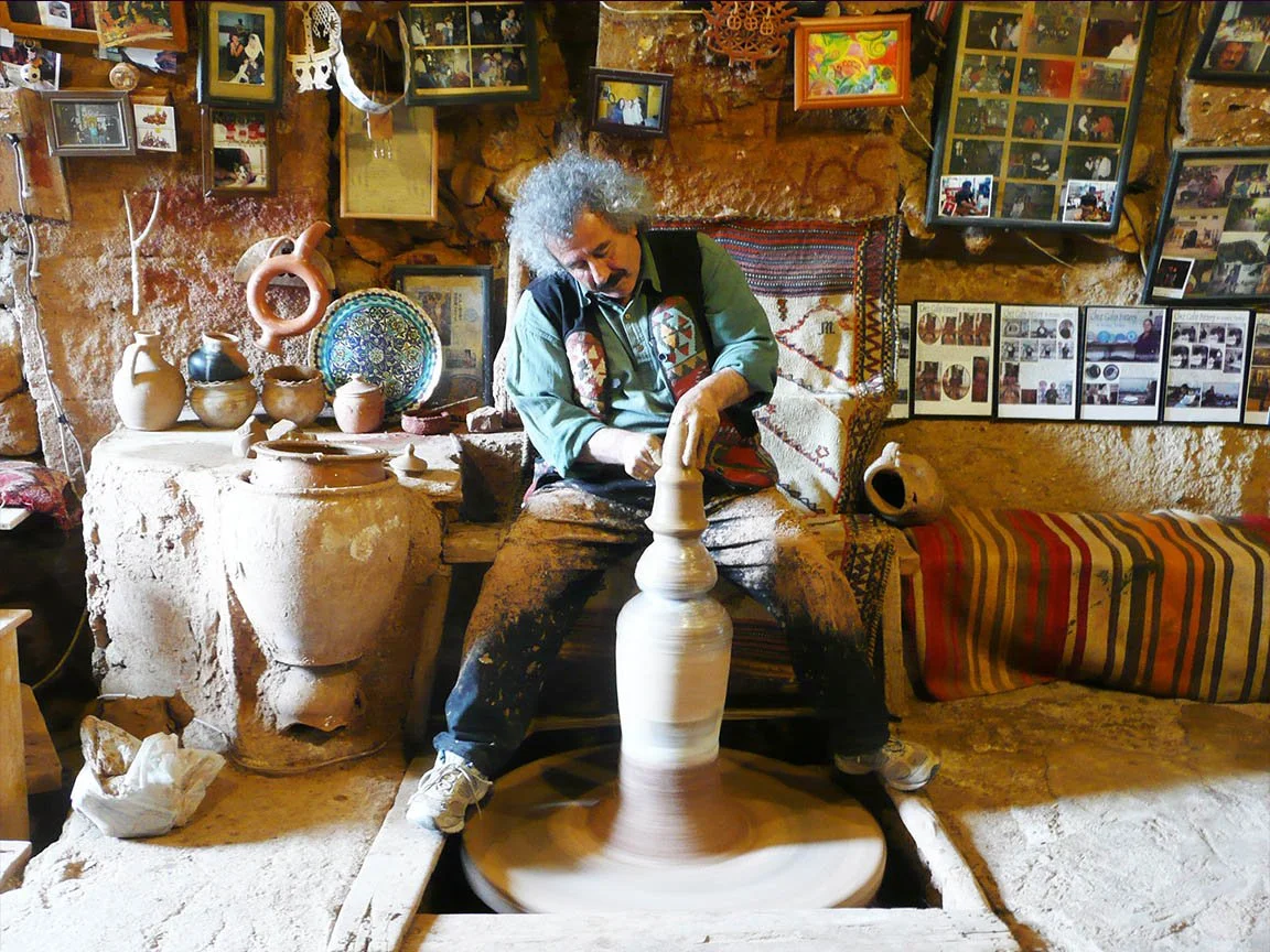 A skilled artisan shaping clay on a traditional potter's wheel in Cappadocia, surrounded by various handcrafted pottery pieces in vibrant colors and intricate designs. The workshop is filled with tools and materials, and the artisan’s focused express