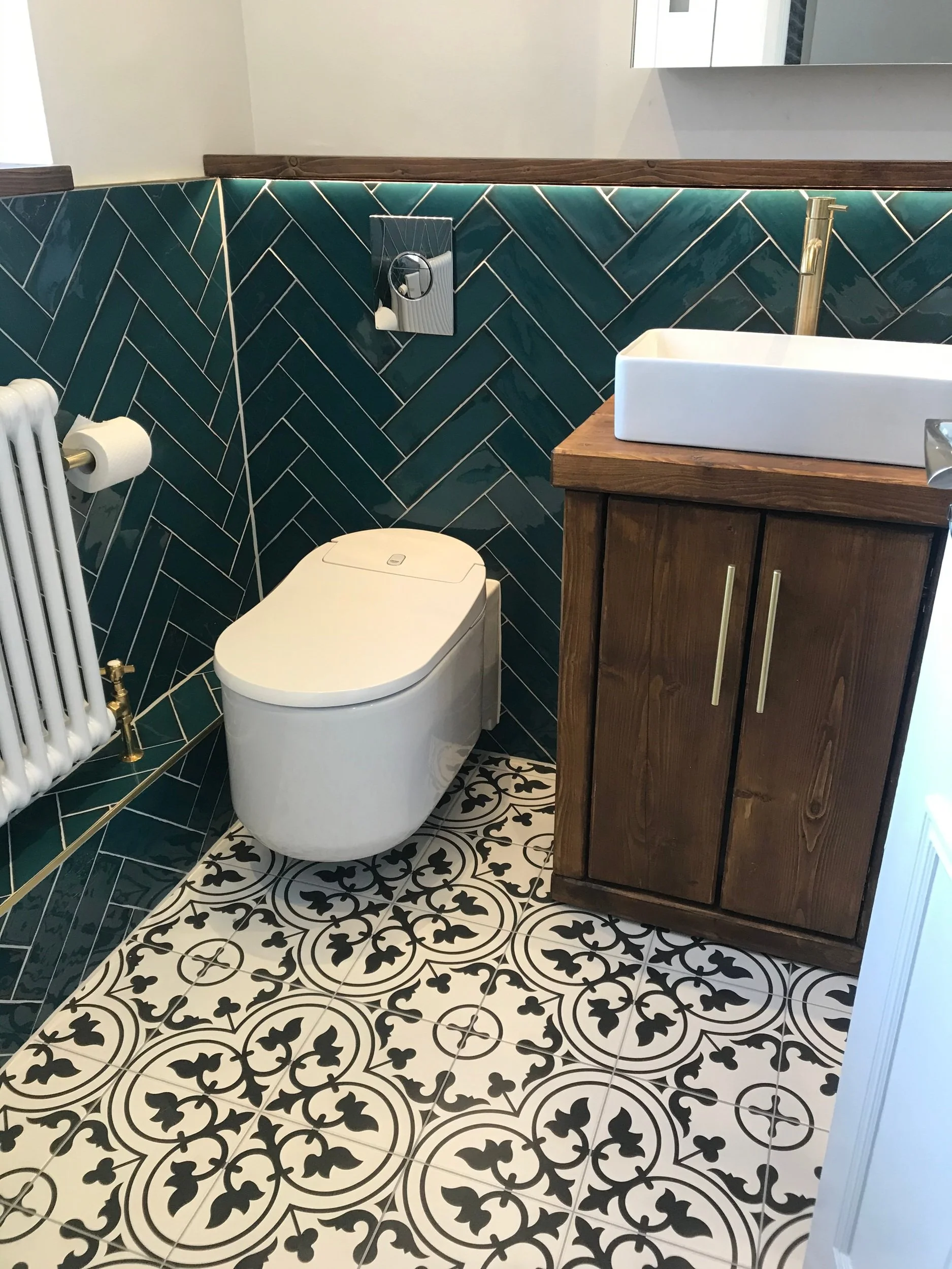 Modern bathroom with geometric patterned black and white floor tiles, teal herringbone tiled wall, white toilet, copper flush button, white sink with wooden cabinet, and gold fixtures.
