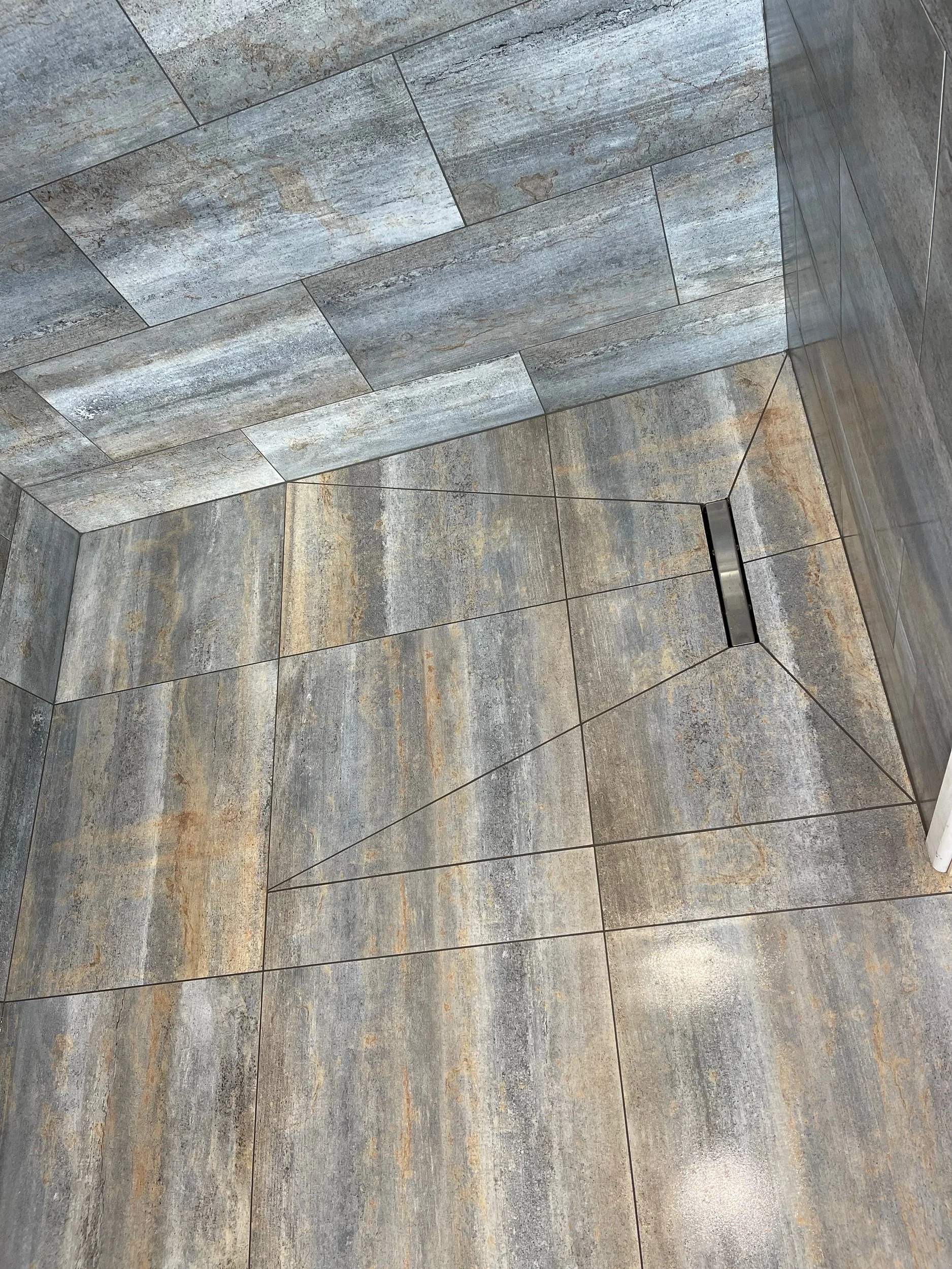 A corner of a tiled shower floor with gray, brown, and beige stones, a drain cover in the center, and tiled walls.