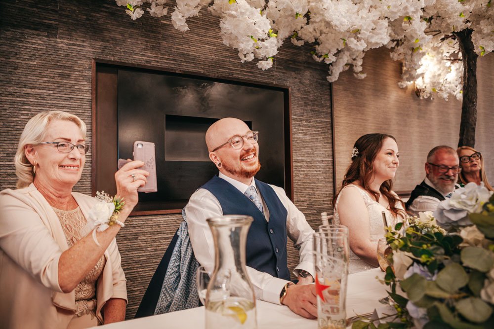 bride and groom smile during speeches as mother records on her phone