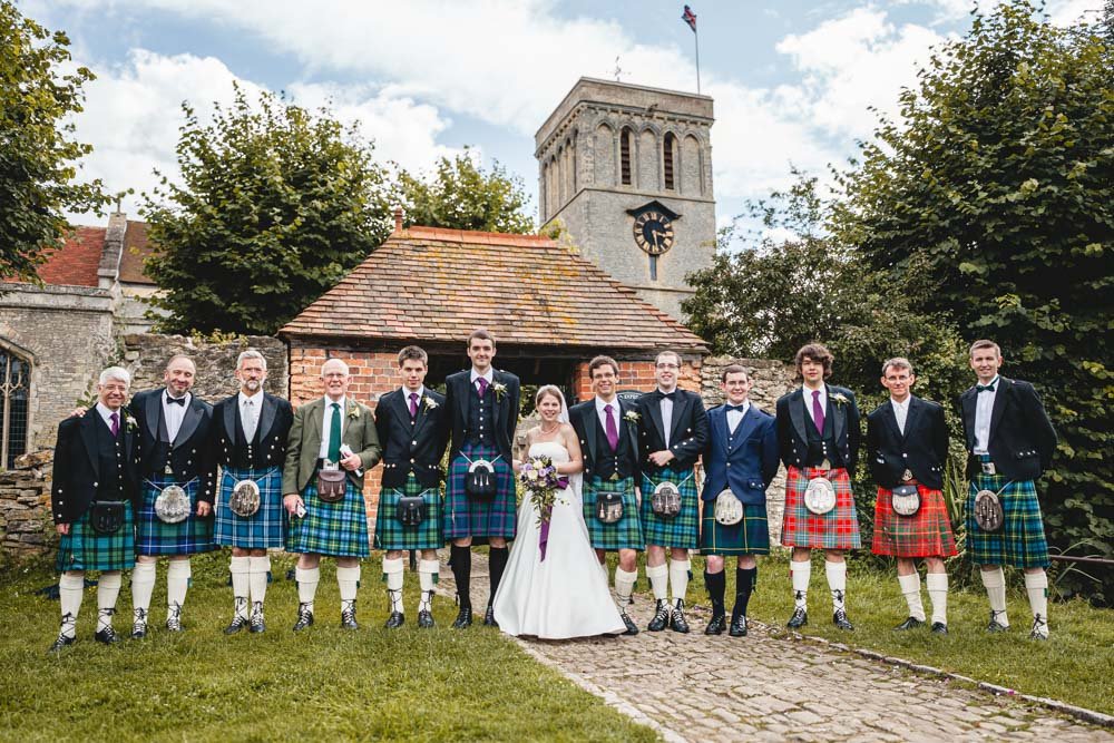 A dozen men in kilts stand with bride in front of Haddenham church in Oxfordshire