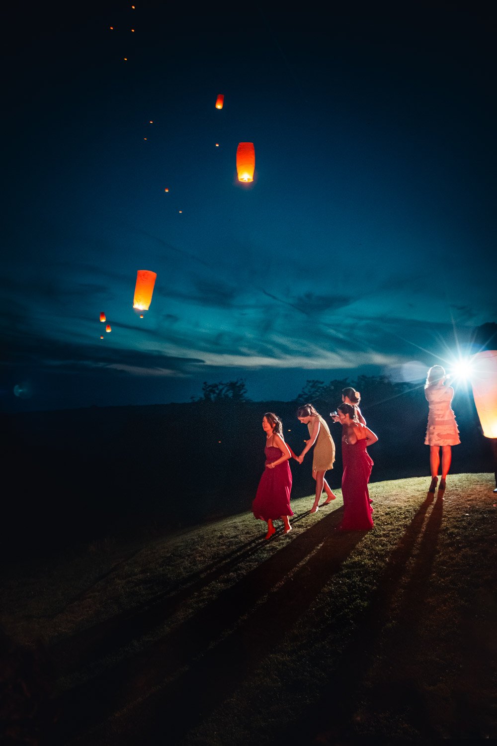 bridesmaids walk on a hill at night, with paper lanterns floating behind