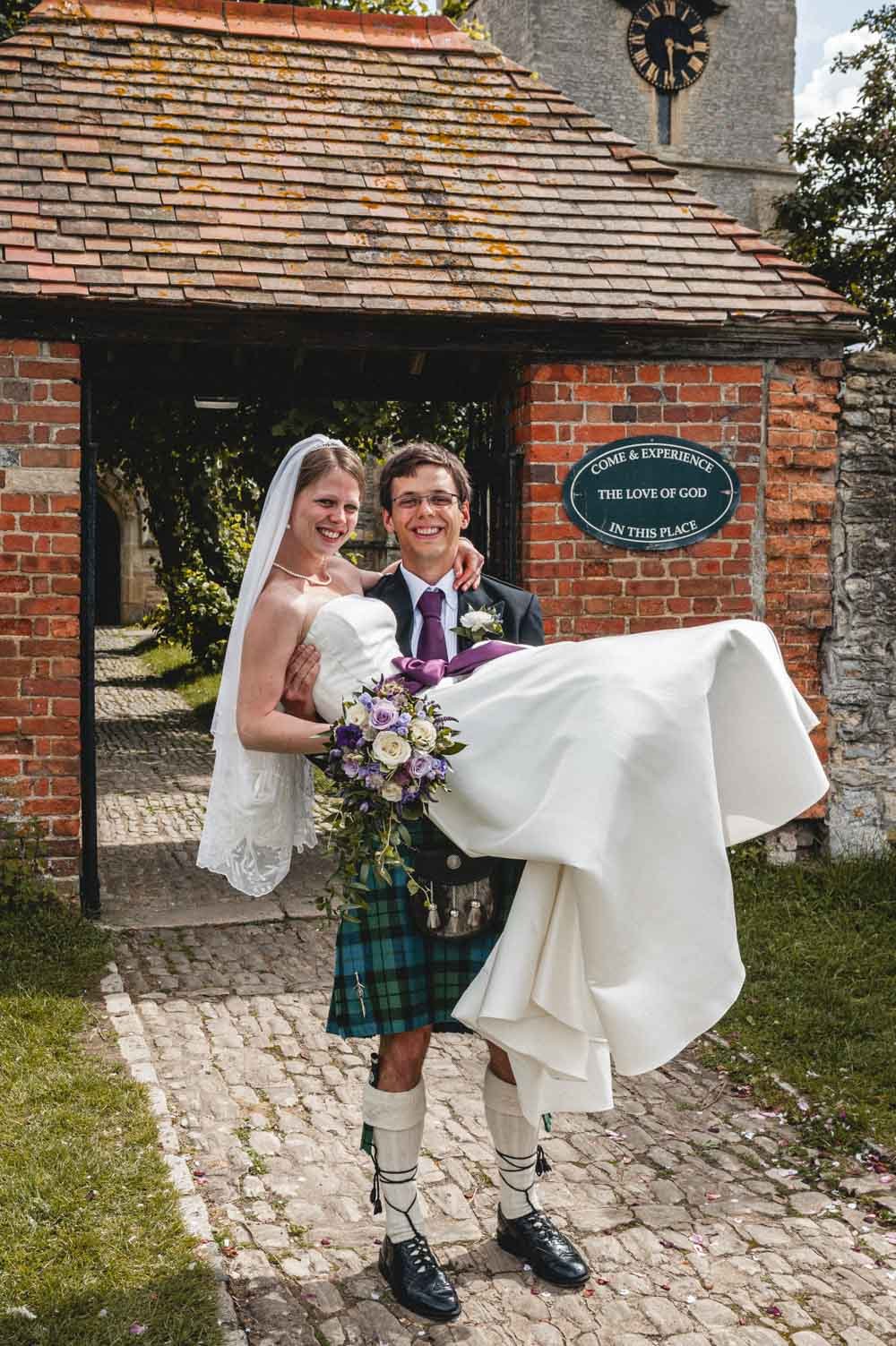 groom carries bride through lychgate outside church
