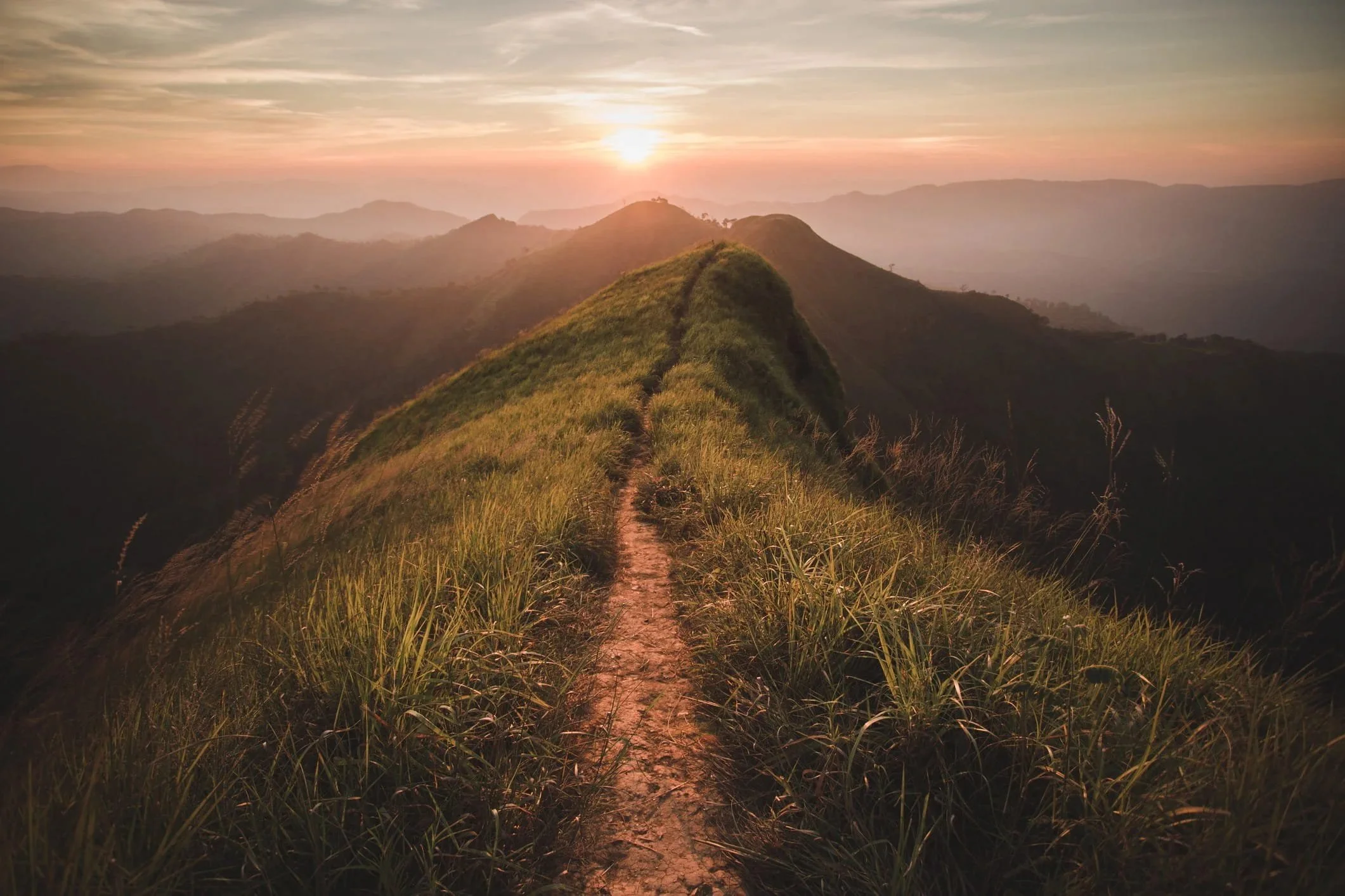 A dirt trail runs along the top of a grassy mountain ridge at sunset, with rolling hills and a colorful sky in the background.