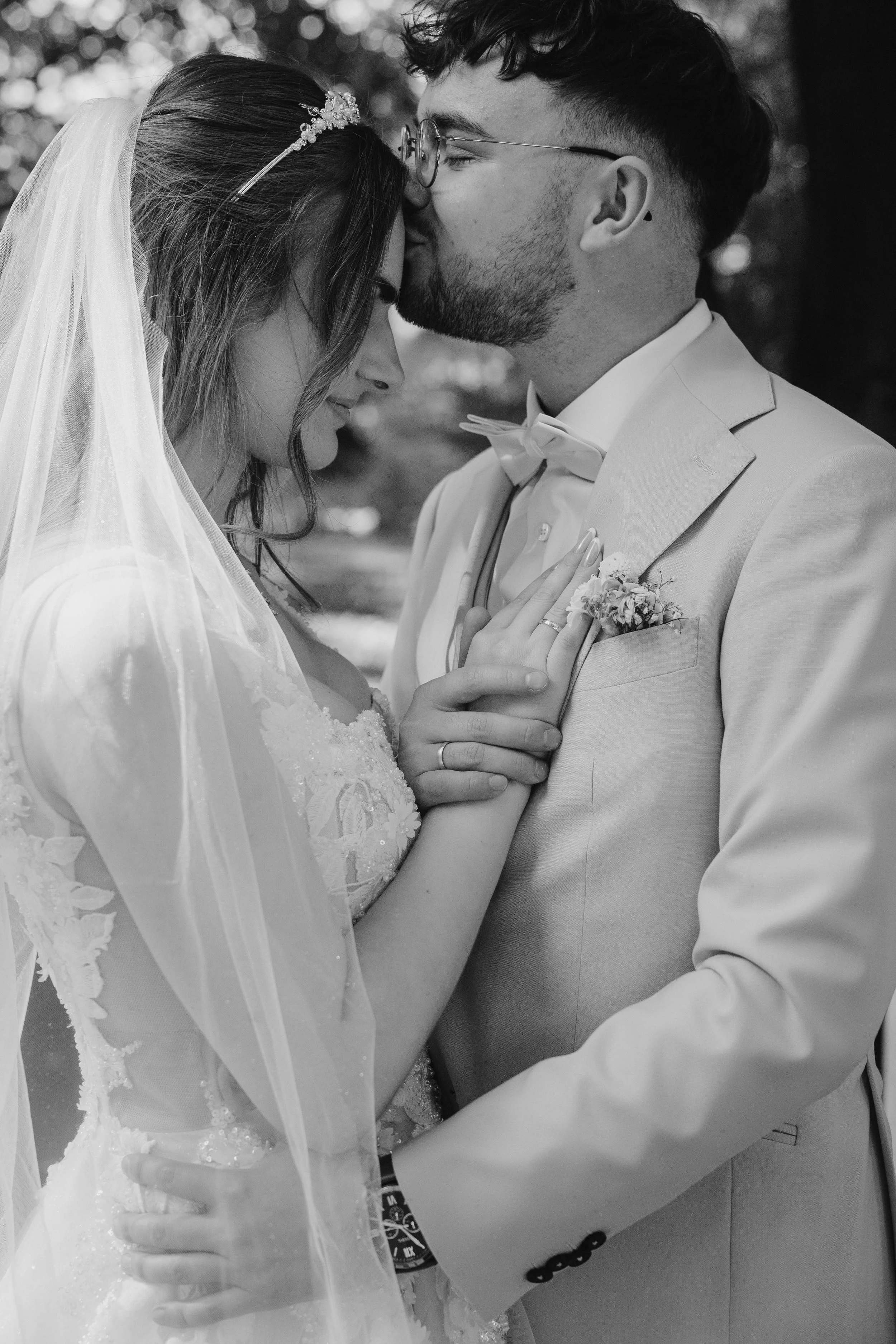 Black and white photo of a bride and groom in an intimate embrace, with their foreheads touching, outdoors, with the bride wearing a wedding dress and veil, and the groom in a light suit and bow tie.