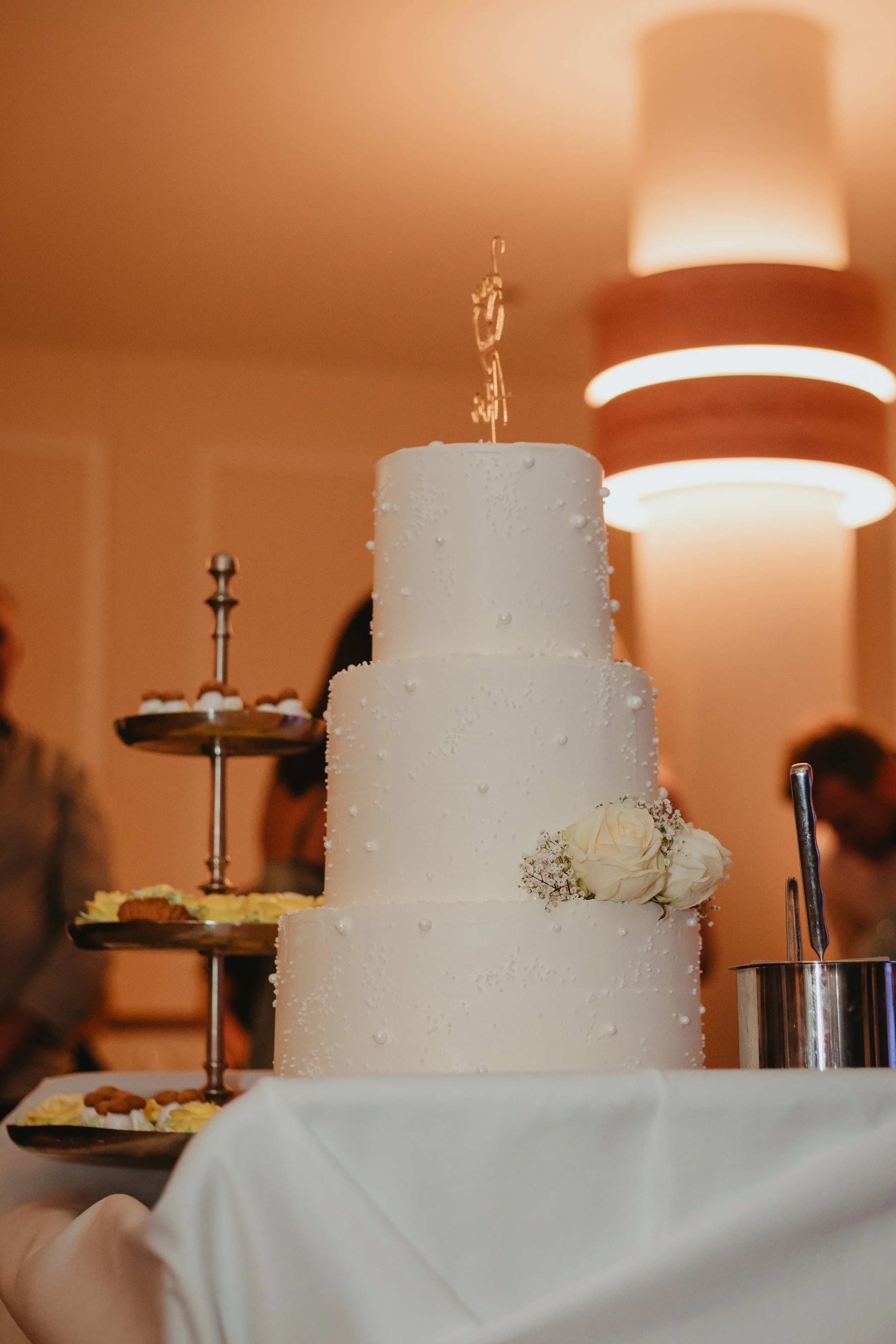 Three-tier white wedding cake with pearl-like decorations and white roses, topped with a decorative cake topper, on a table with a white tablecloth at a wedding reception.