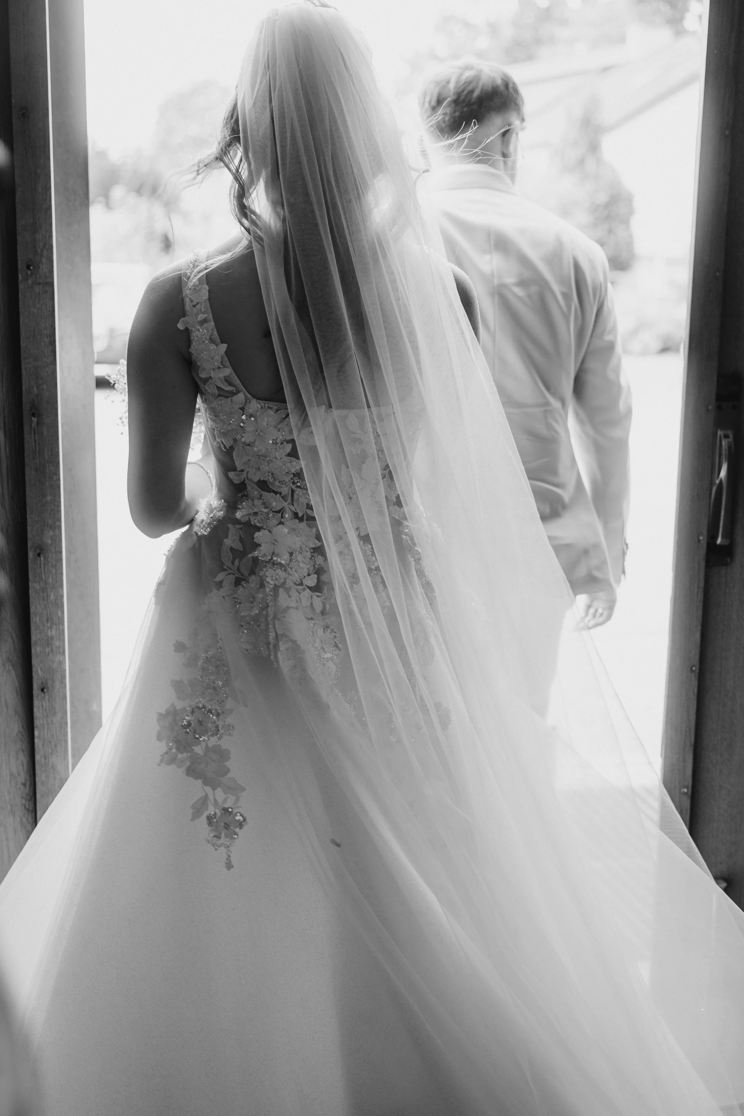 Black and white photo of a bride in a wedding dress with a veil, standing with her back to the camera, looking out a doorway, with a man in a shirt beside her.