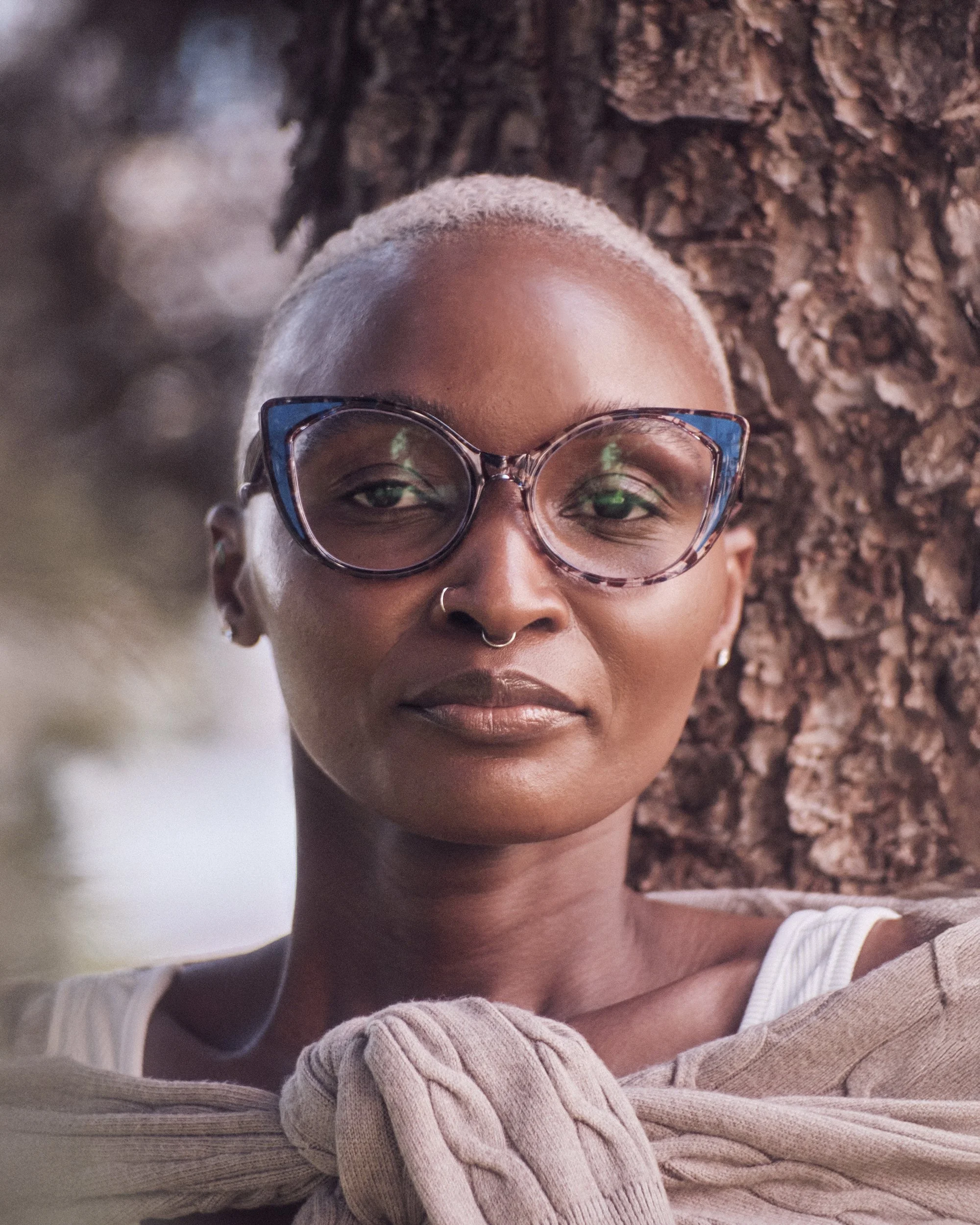 A woman with short platinum blonde hair wearing large, colorful glasses, standing outdoors near a tree trunk.