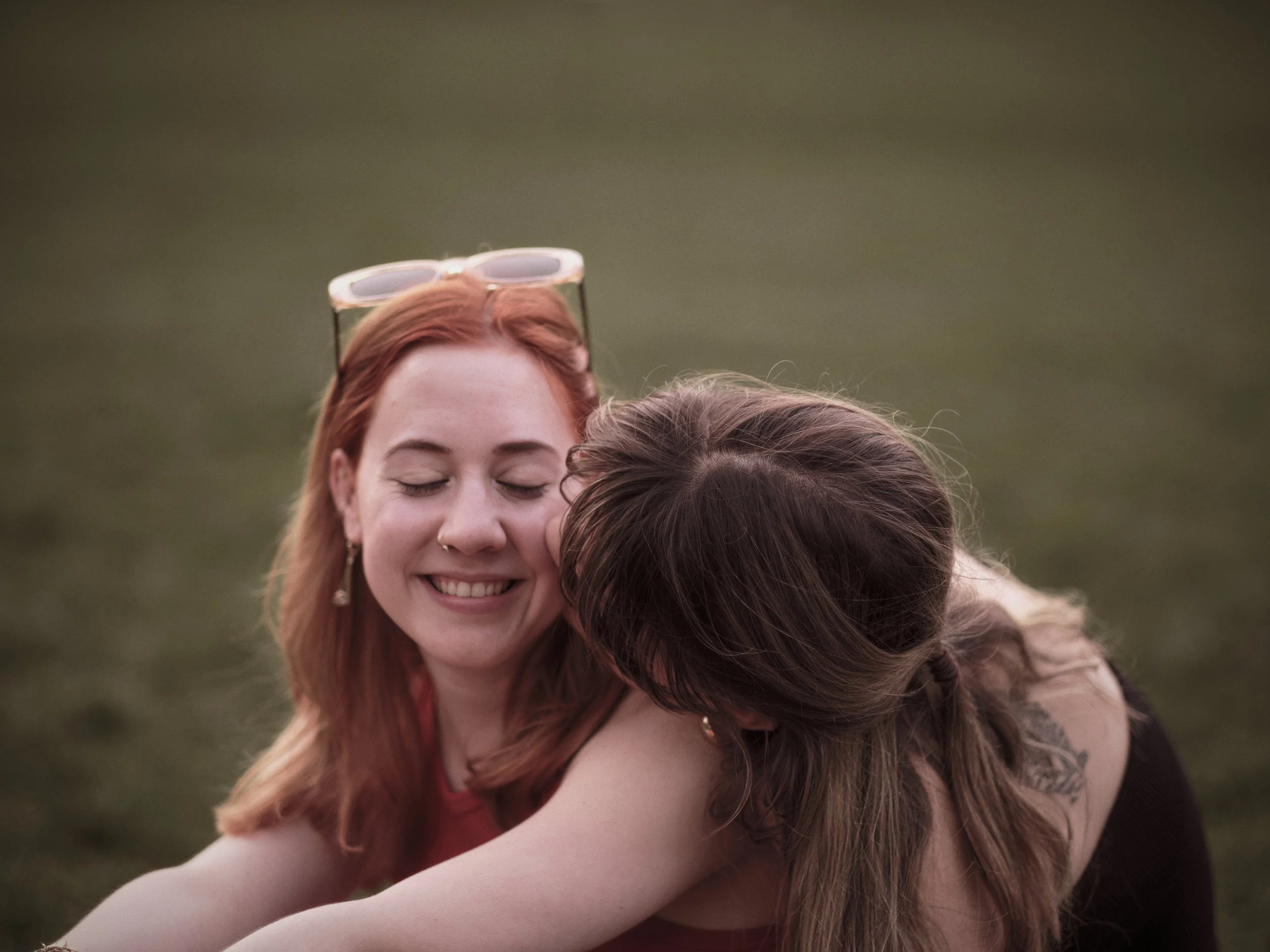 Two women enjoying a hug outdoors on grass, one with red hair, sunglasses on her head, and a nose ring, smiling with eyes closed, while the other with brown hair leans in, partially obscured.