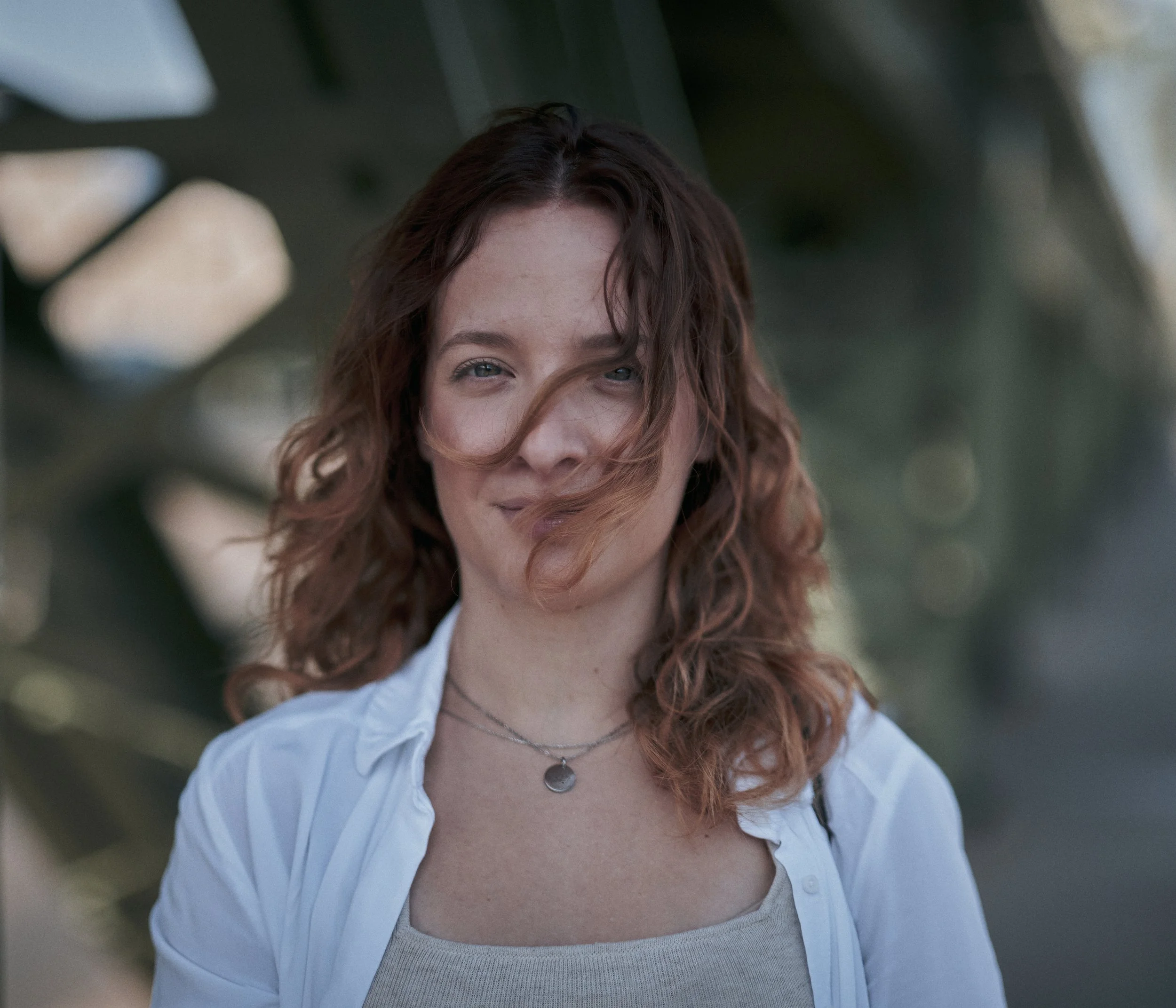Young woman with curly hair smiling outdoors, wearing a white shirt and layered necklaces.