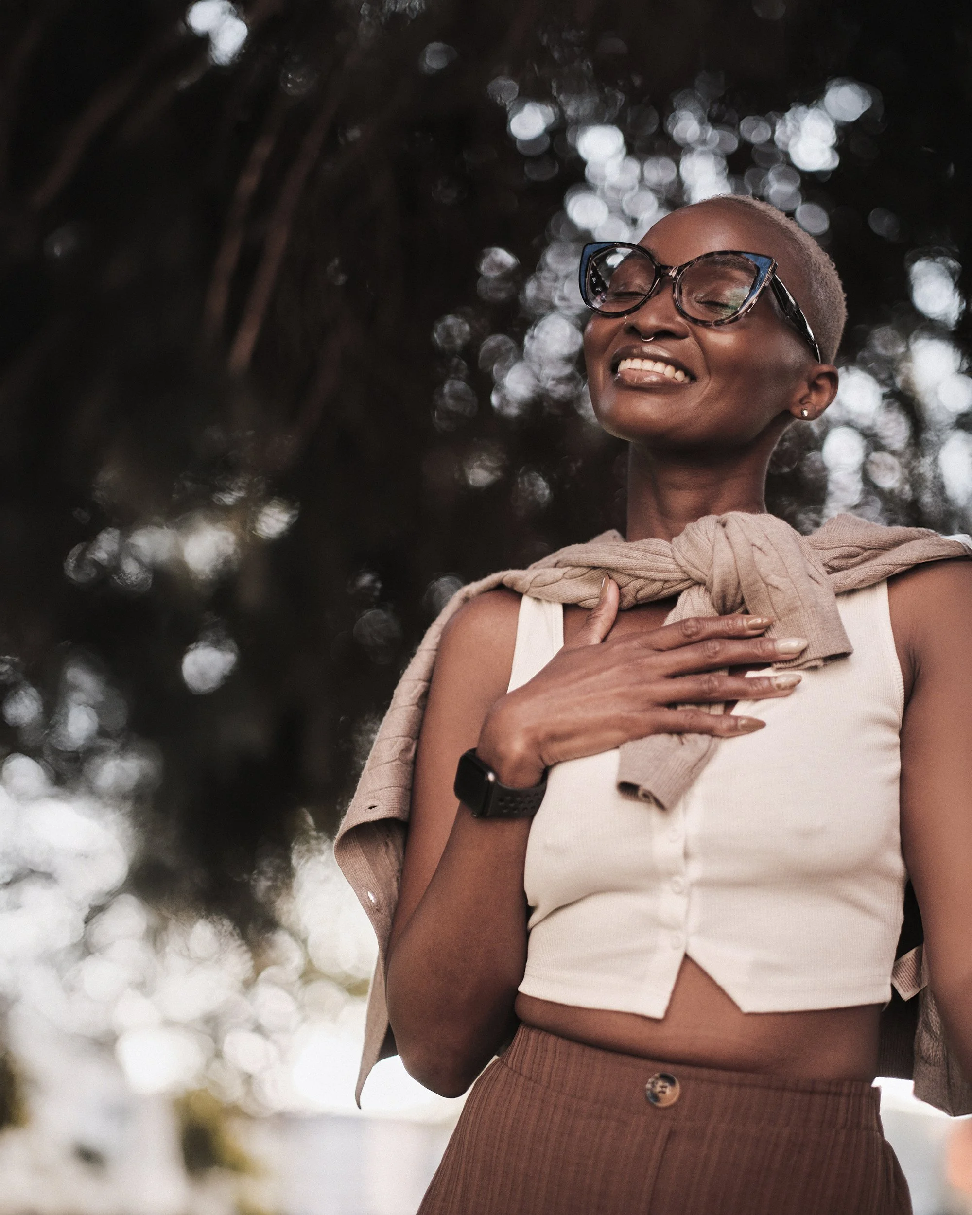 A woman smiling with her eyes closed, wearing glasses, a white tank top, and a beige sweater draped over her shoulder, standing outdoors with blurred trees and lights in the background.