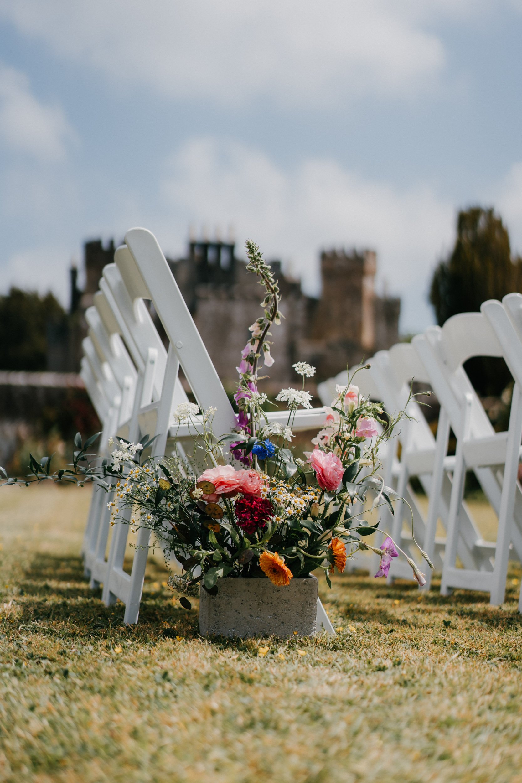 White folding chairs arranged outdoors with a vibrant floral arrangement on the grass, castle in the background.