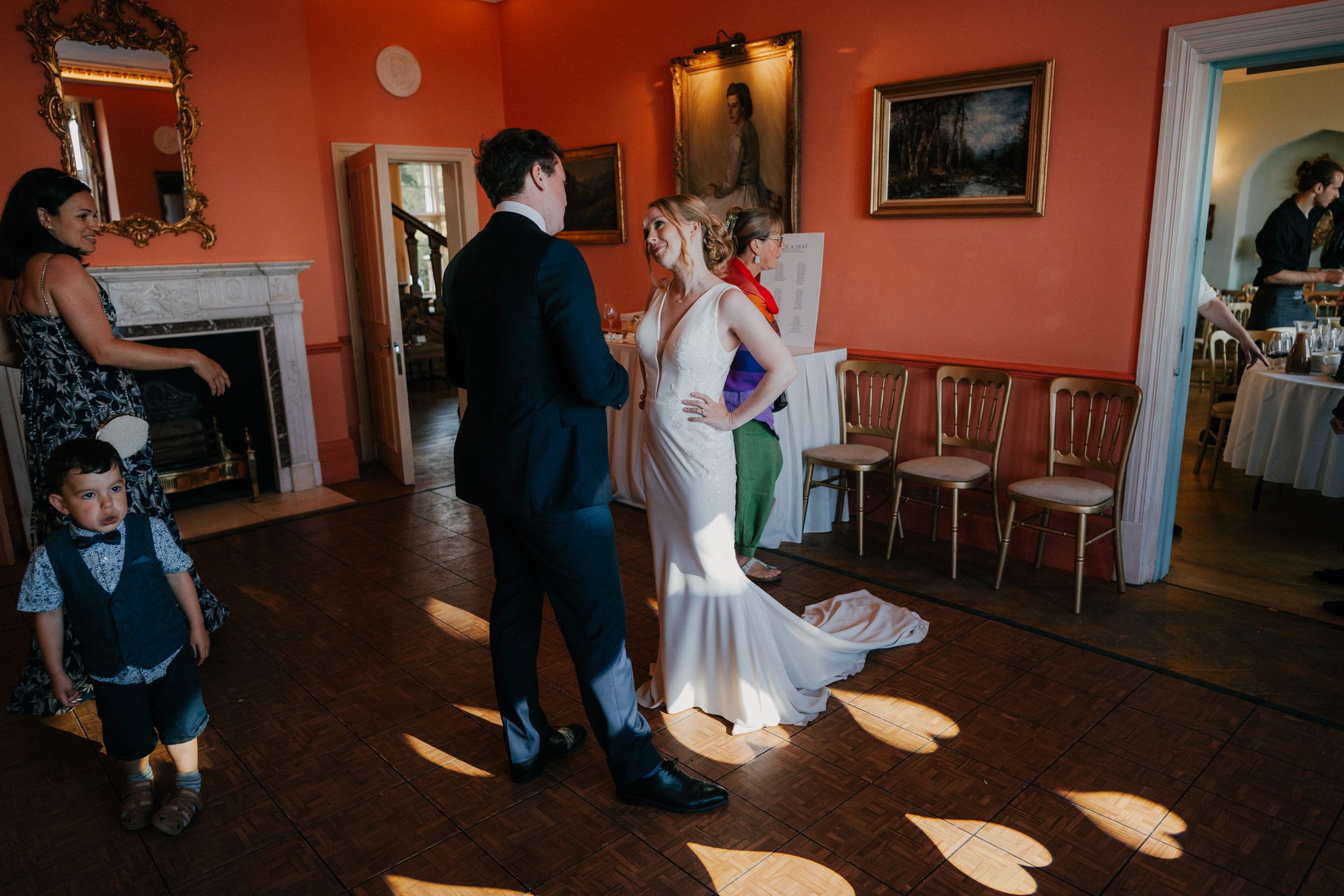 Guests at a wedding reception in a room with orange walls, featuring a bride in a white dress conversing with a man in a suit. Other attendees, including a child, are present. Framed artwork and mirrors adorn the walls, and there is a table with a ch