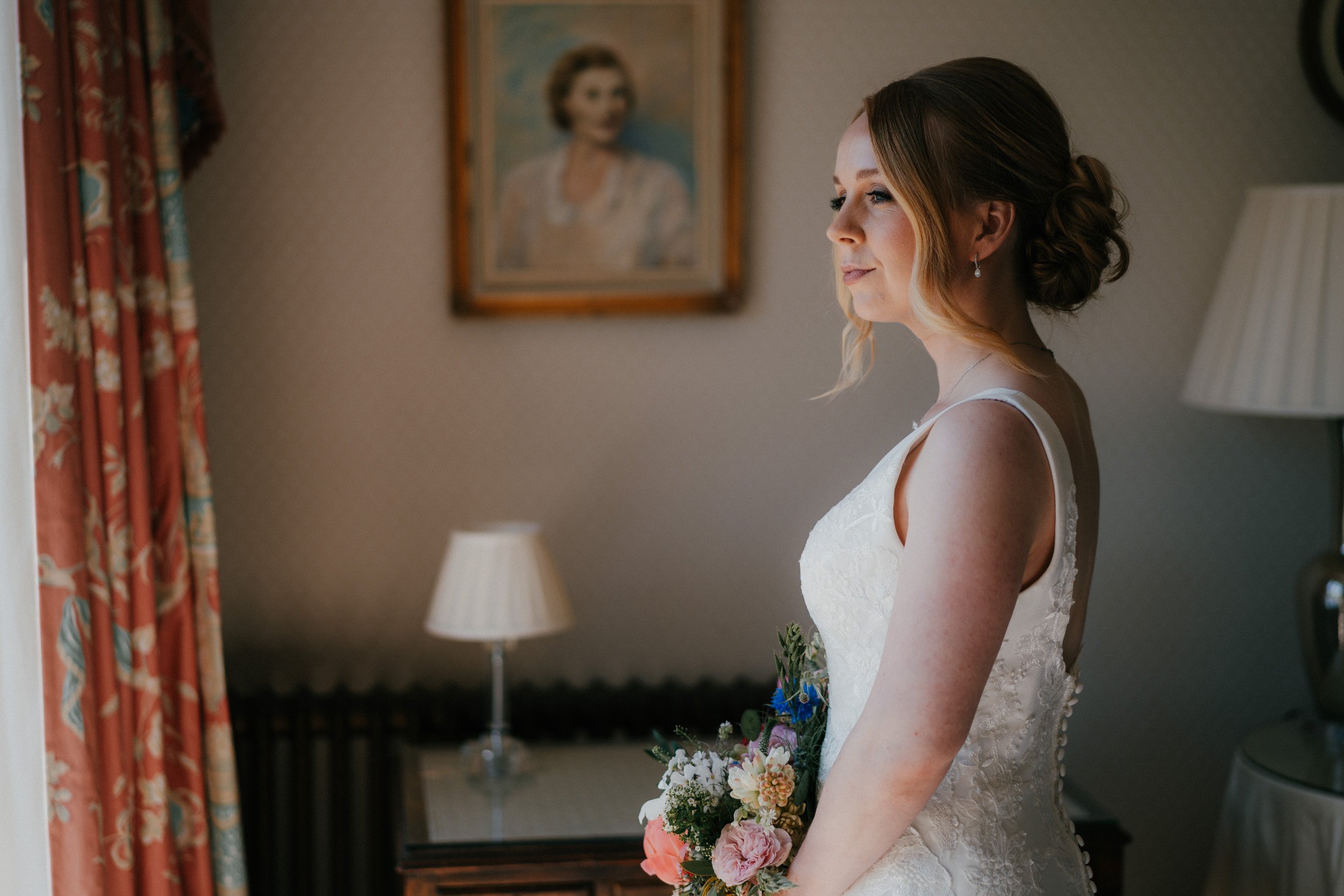Bride holding bouquet, standing by window in a room with floral curtains and table lamp.