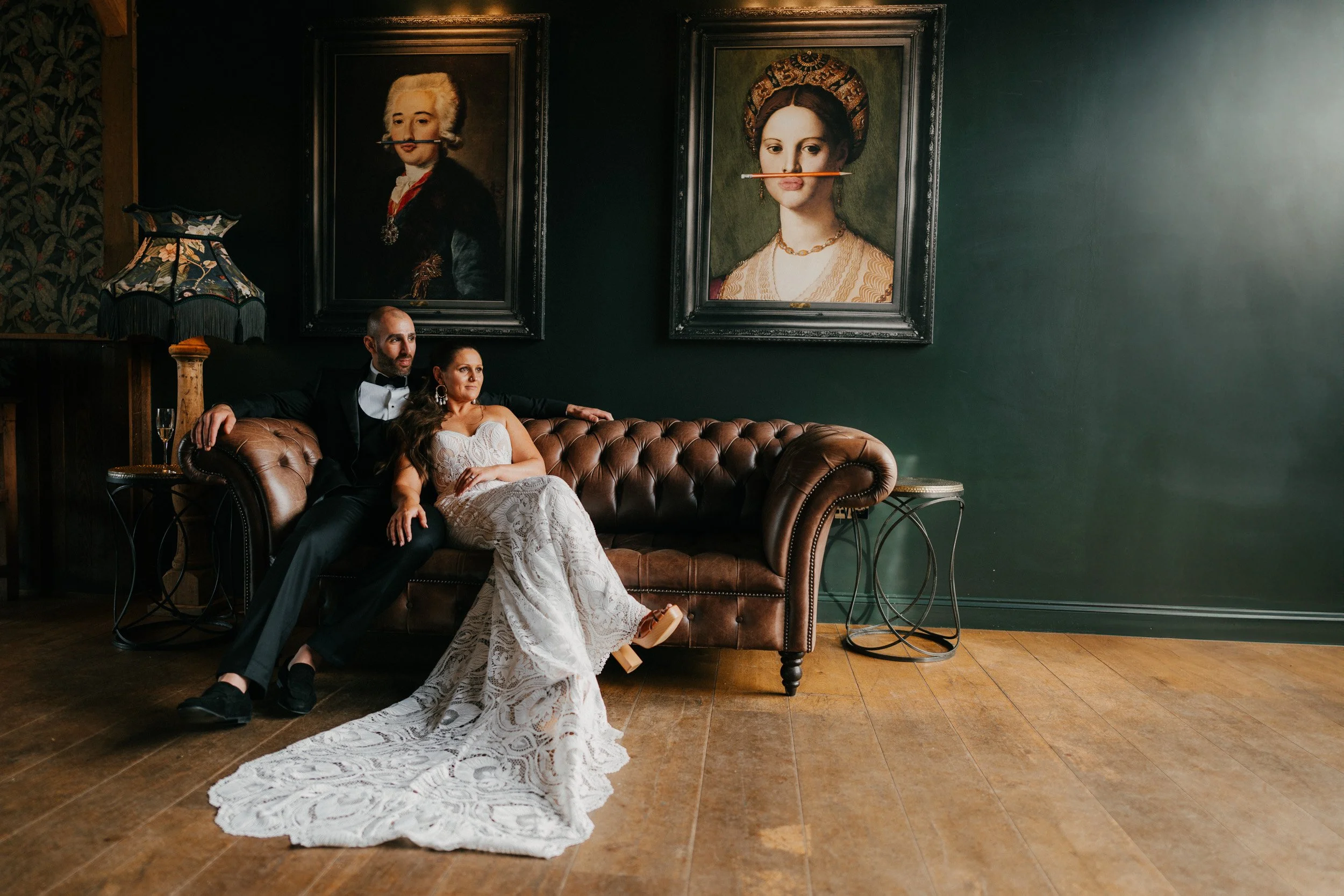 A couple dressed in formal attire, sitting on a leather couch in an elegant room with framed portraits and ornate décor.