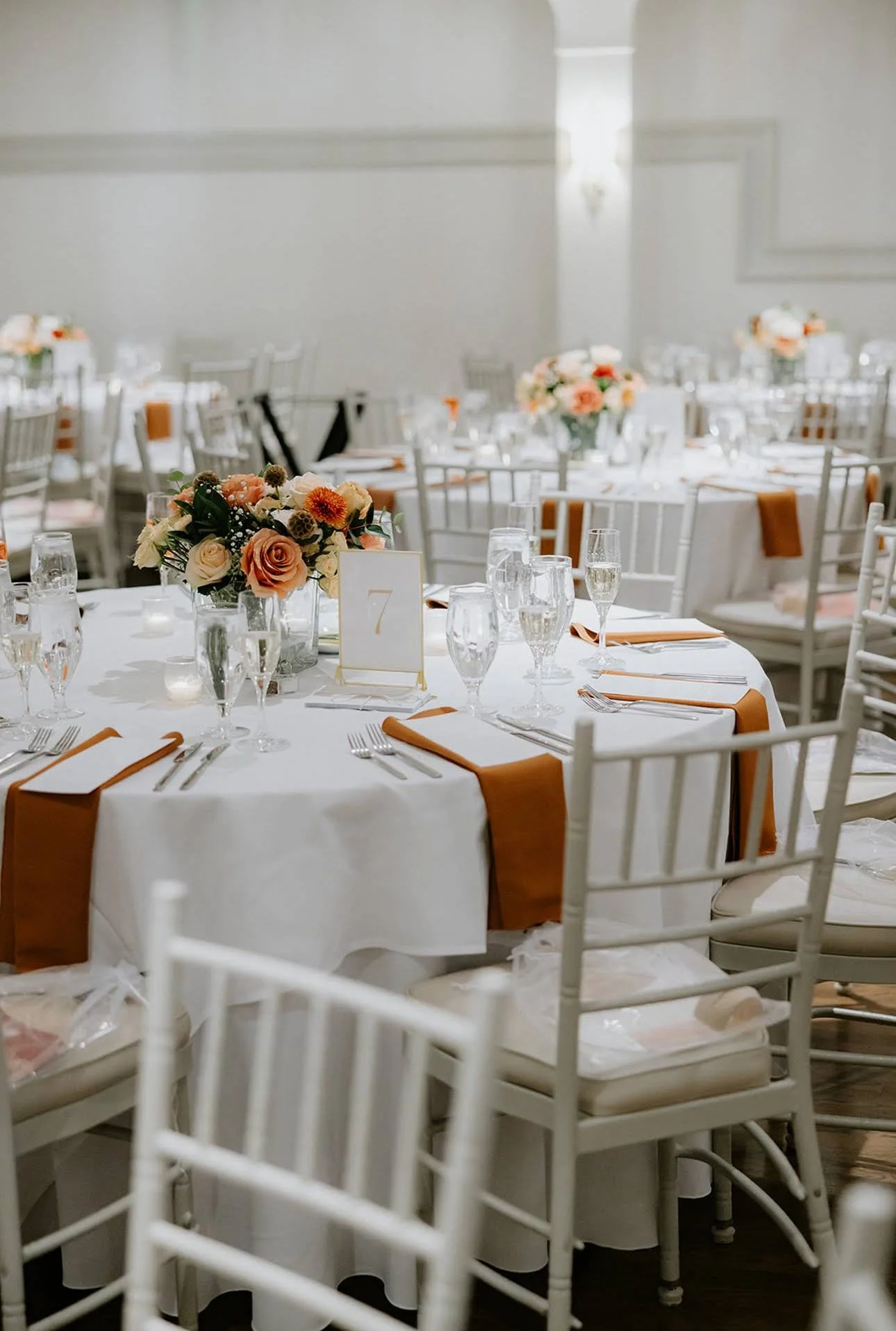 Formal event setup with a round table decorated with a floral centerpiece, multiple champagne flutes, water glasses, cutlery, and napkins, in a white-themed room with elegant chairs and soft lighting.