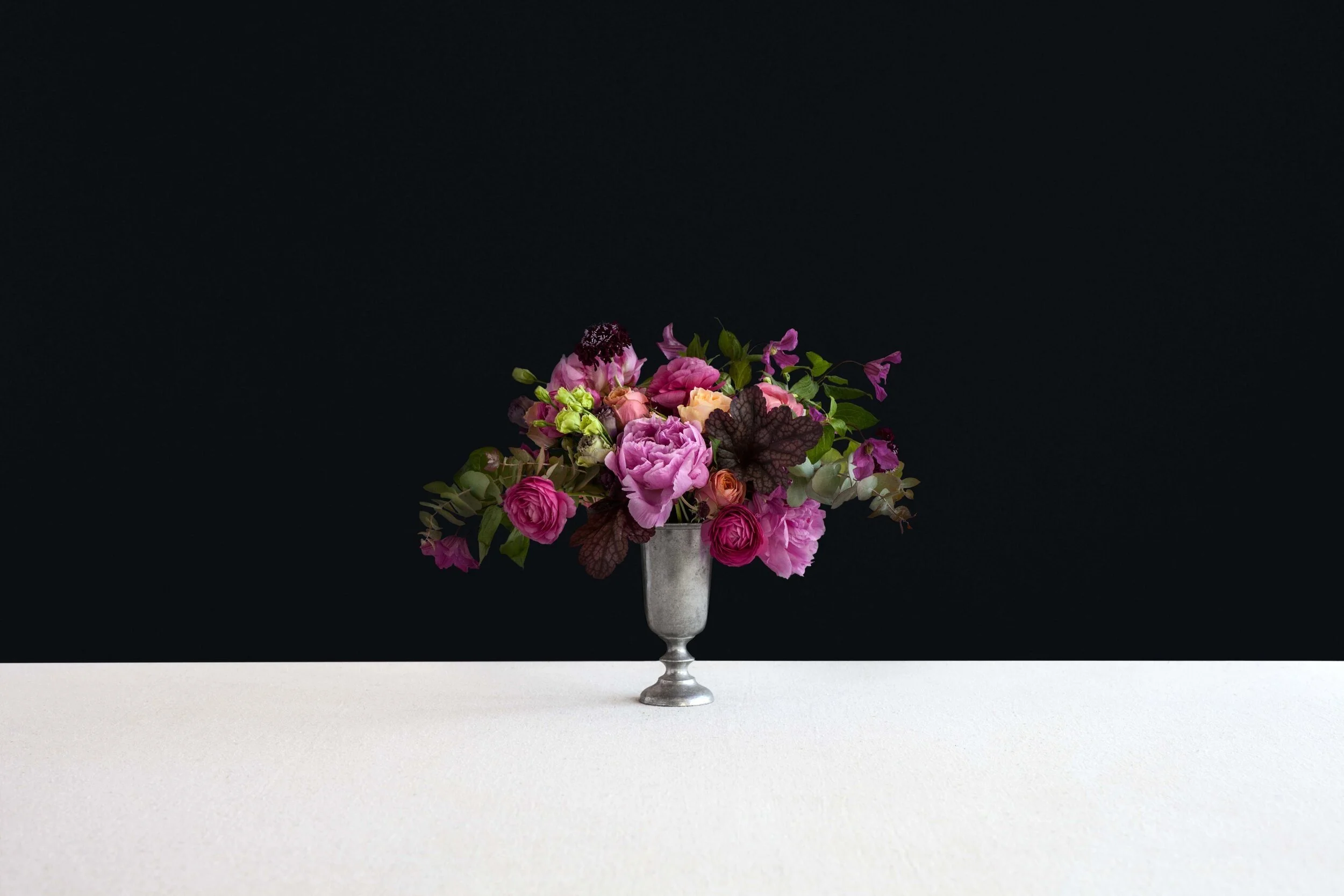 A bouquet of pink, purple, and green flowers in a silver vase on a white surface against a black background.