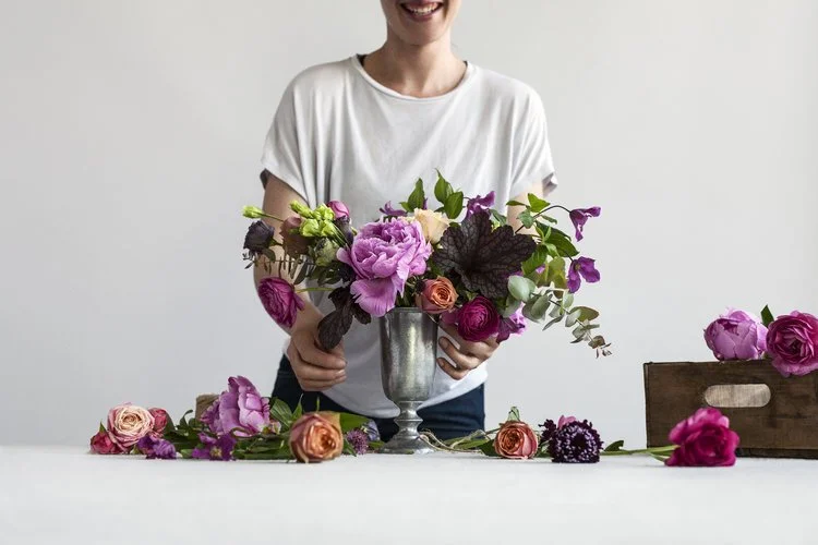 A person holding a silver vase filled with pink, purple, and peach flowers, with additional flowers on a white table and a wooden box on the right side.