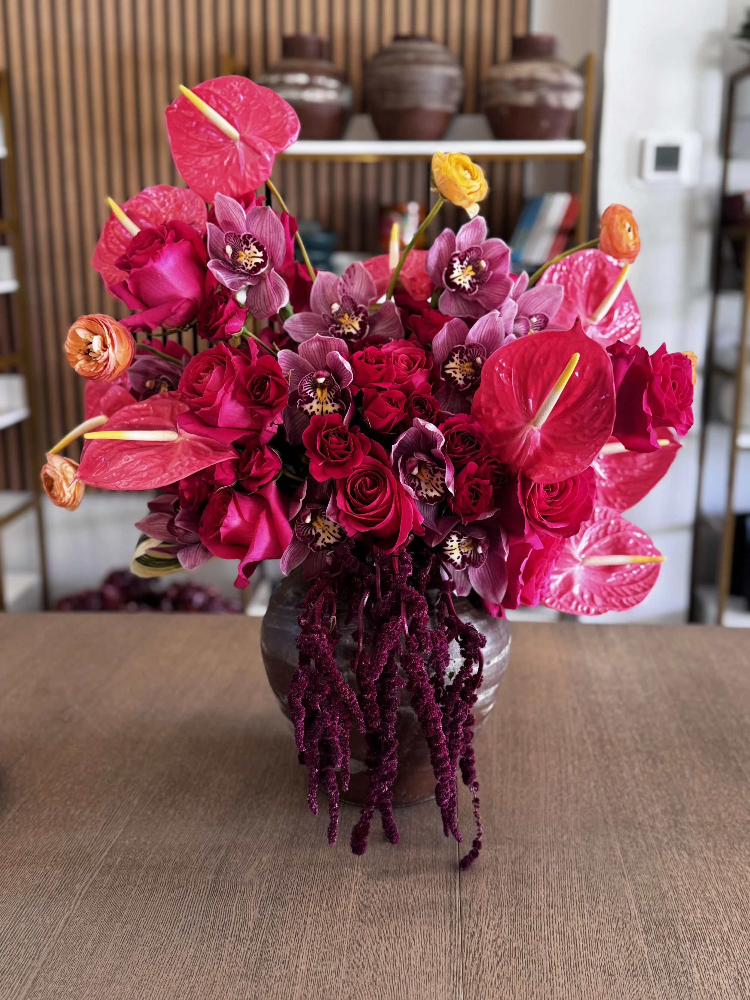A vibrant flower arrangement in a dark ceramic vase featuring pink roses, pink anthuriums, purple orchids, and purple hanging amaranthus. The background shows a wooden shelf with decorative vases and books.