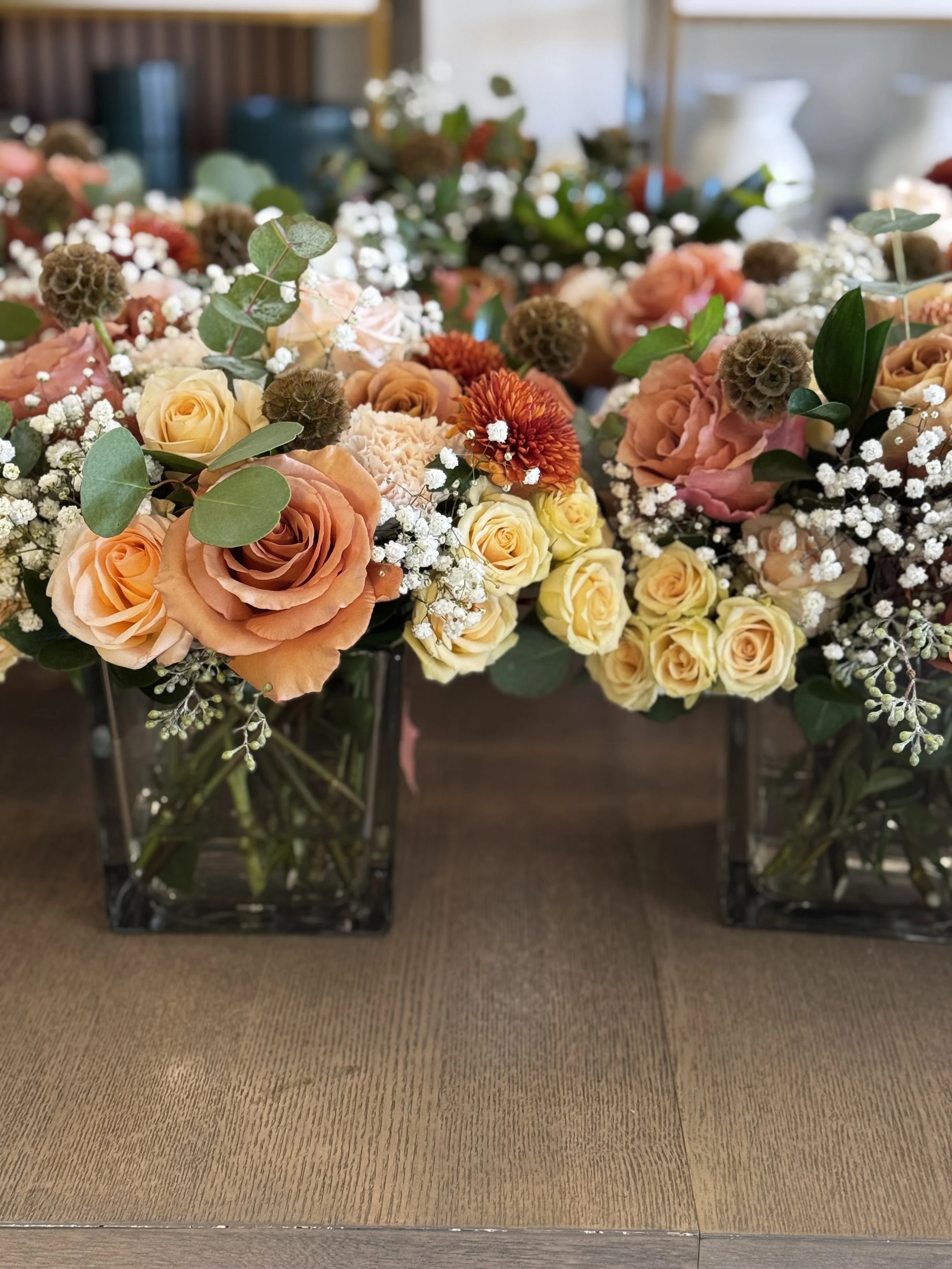 Two glass vases filled with peach, cream, and blush roses, white baby's breath, eucalyptus leaves, and other small flowers sitting on a wooden surface.