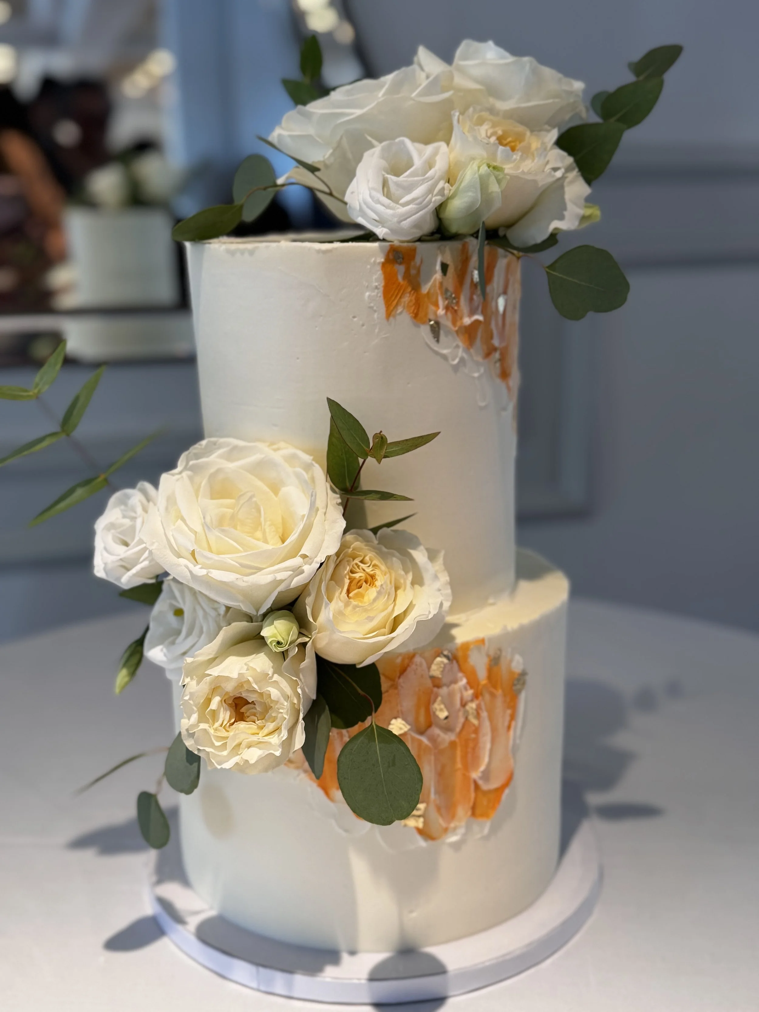 Two-tier white wedding cake decorated with white roses, cream-colored flowers, and green leaves, with orange decorative accents