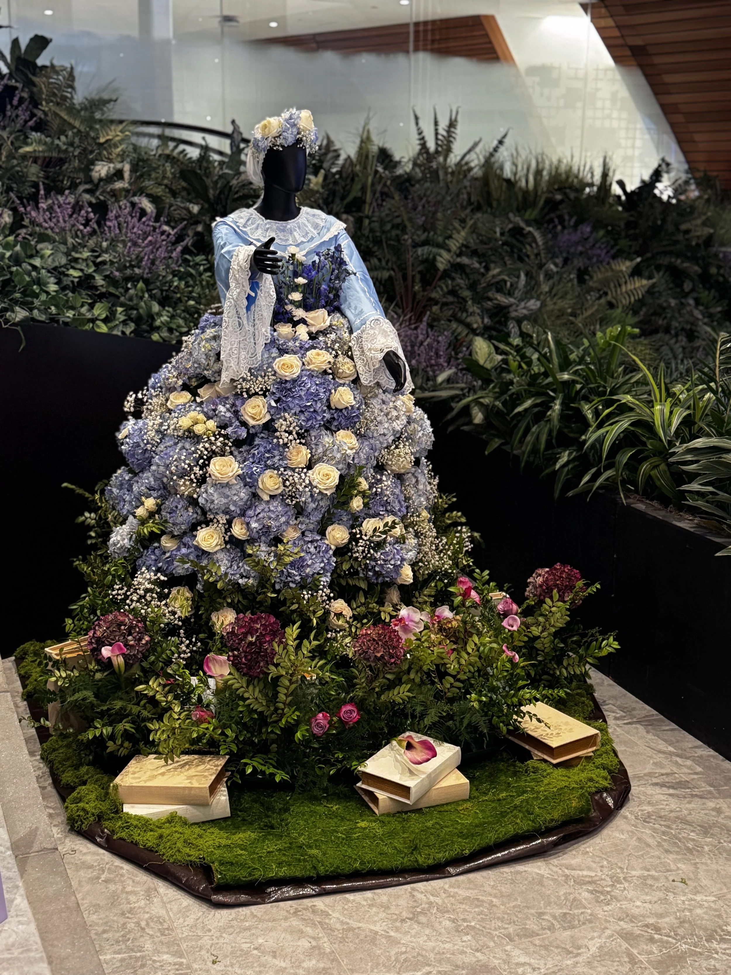 Display of a mannequin dressed in a blue vintage-style dress with lace details, adorned with a floral headpiece, surrounded by a large arrangement of purple and white flowers, with some pink roses and books at the base, set against a lush garden or i
