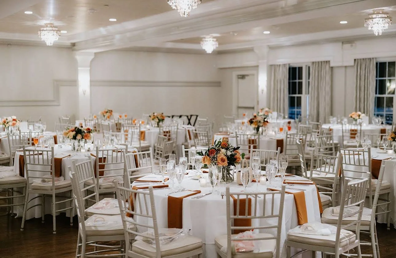 Elegant banquet hall decorated with round tables draped in white tablecloths, adorned with floral centerpieces and set with glassware, plates, and utensils, with chairs arranged around each table and chandeliers hanging from the ceiling.